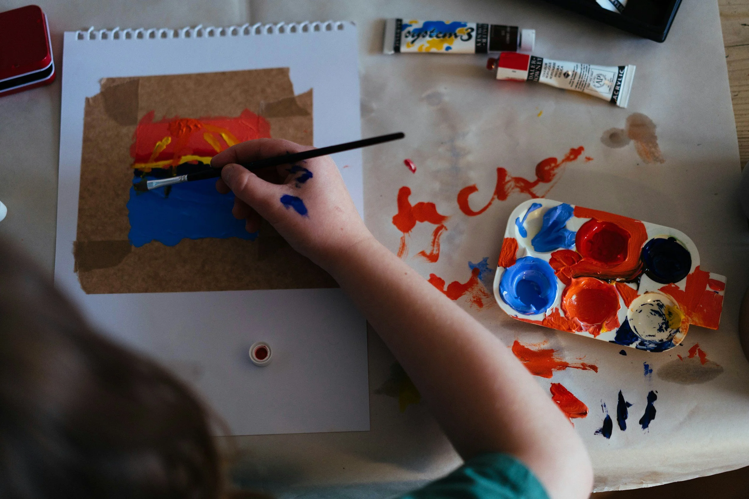 Person painting abstract shapes with red, blue, and yellow paints on paper using a brush, with paint trays and tubes of acrylic paint nearby.
