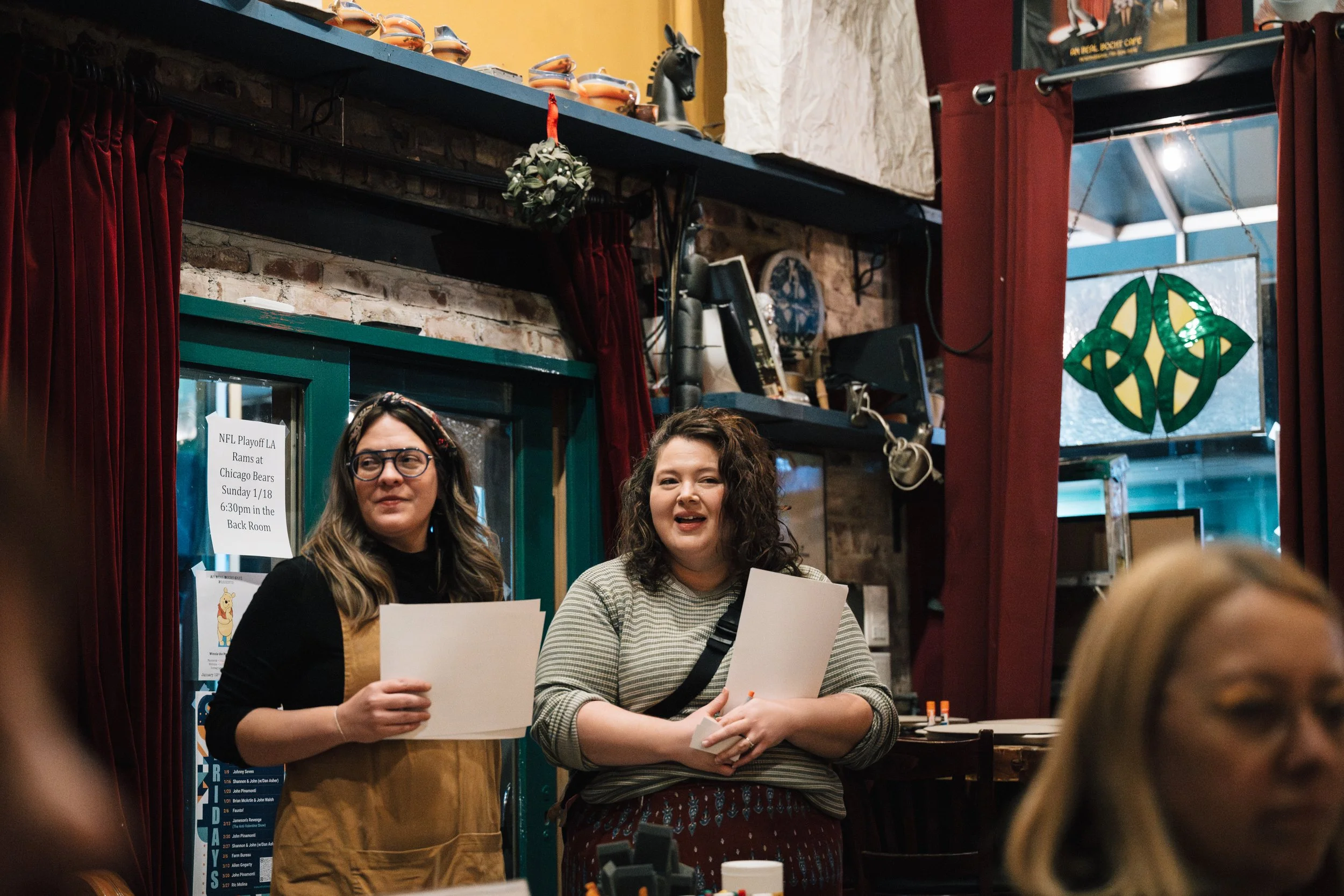 Two women standing behind a table, holding papers, at an indoor event with a brick wall, shelves with decorative items, and a window with a stained glass design in the background.