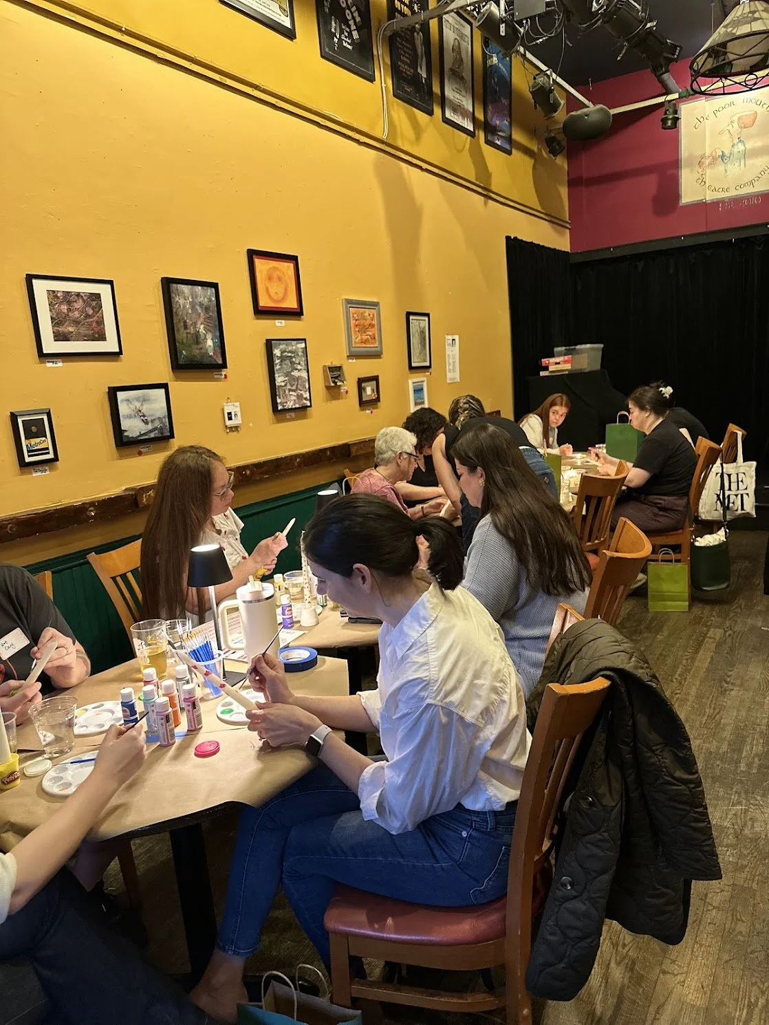 People sitting at a long table painting and customizing items in an art workshop inside a cozy, colorful cafe or art studio. Several framed pictures adorn the yellow wall behind them, and art supplies are on the table.