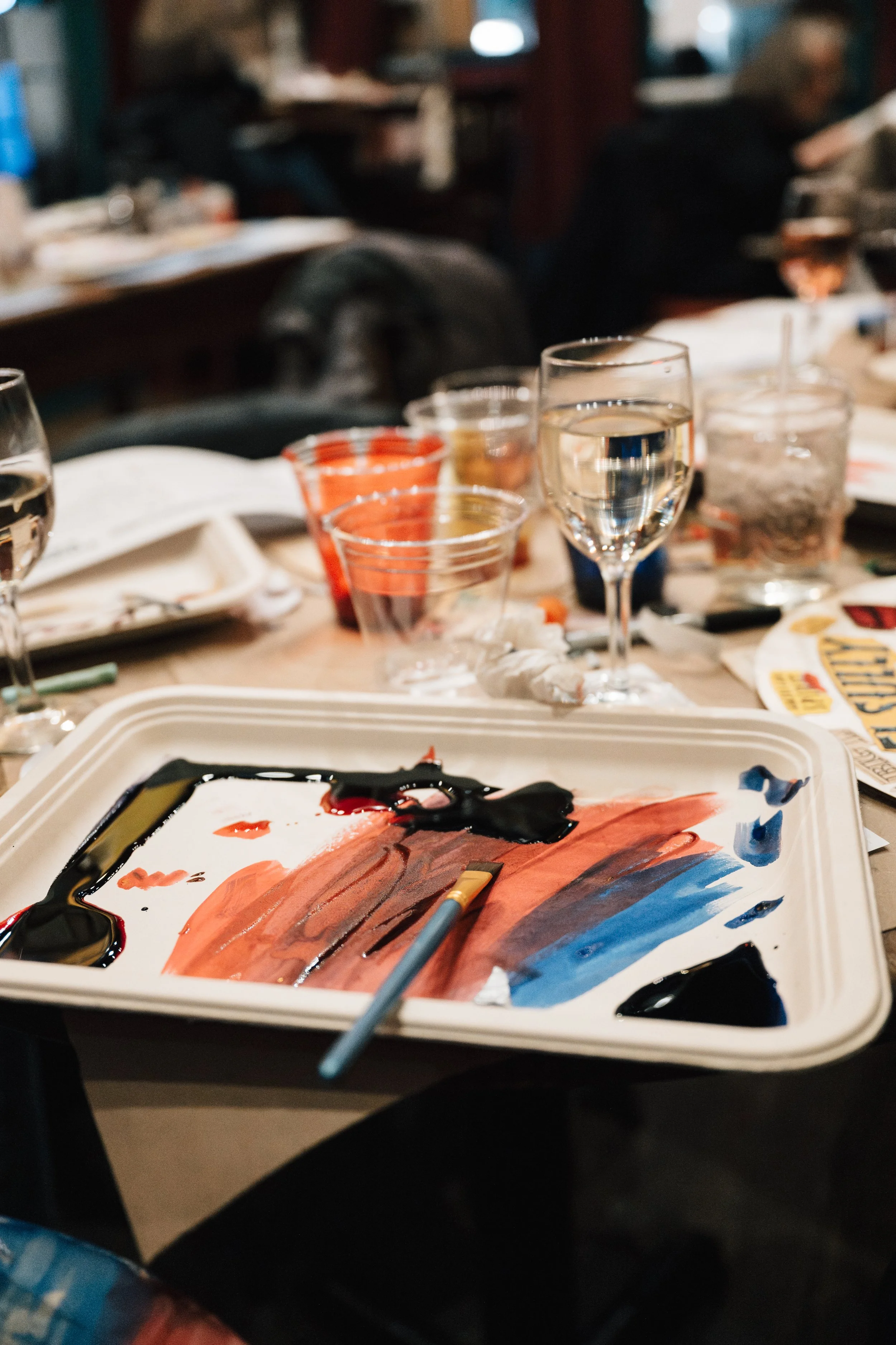 A table with a painting palette, brushes, glasses of water and a glass of white wine. Background shows people sitting and talking in a restaurant or cafe.
