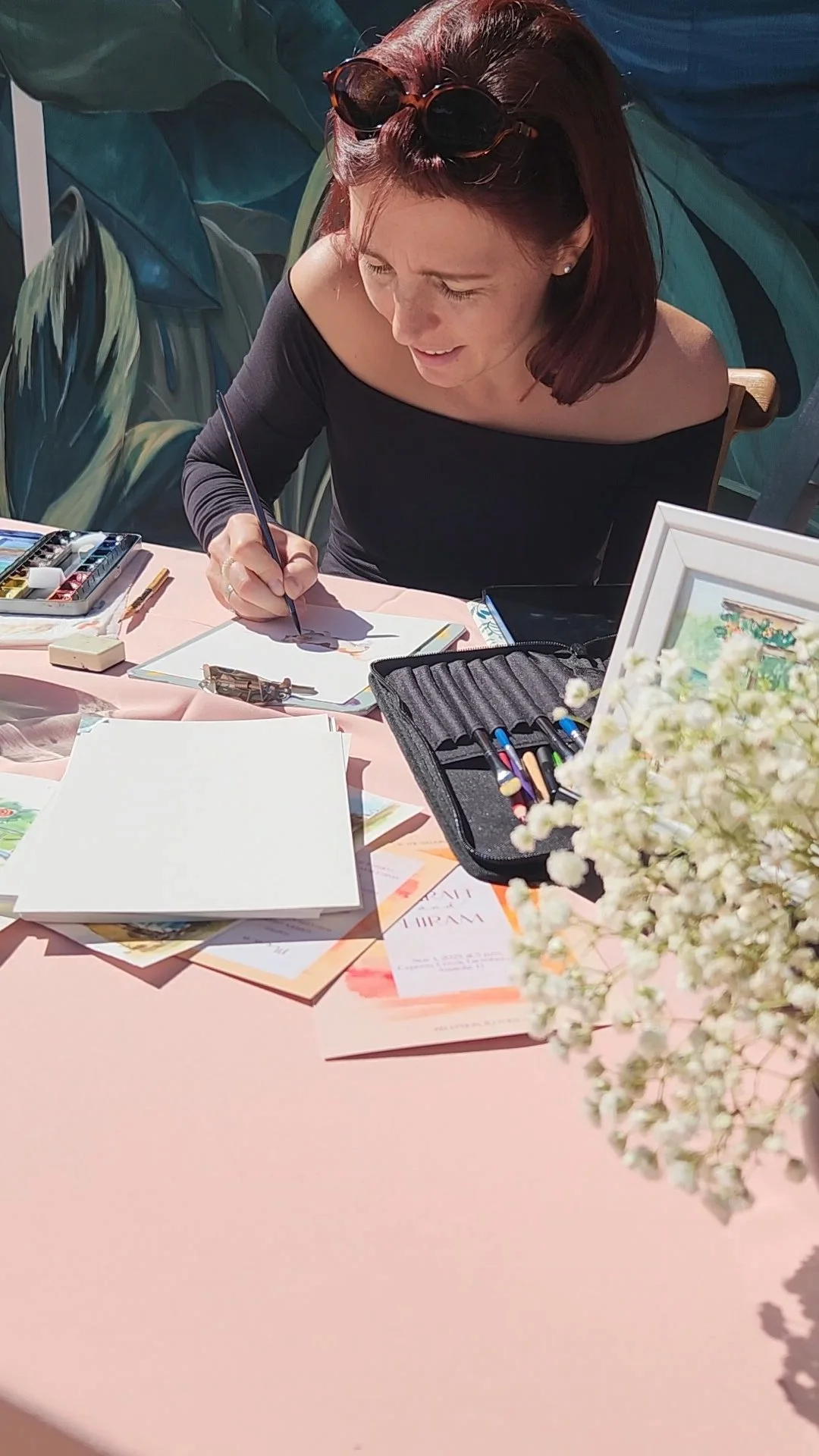 Woman with sunglasses on her head, seated at a pink table, drawing or writing in a notebook surrounded by art supplies and flowers.