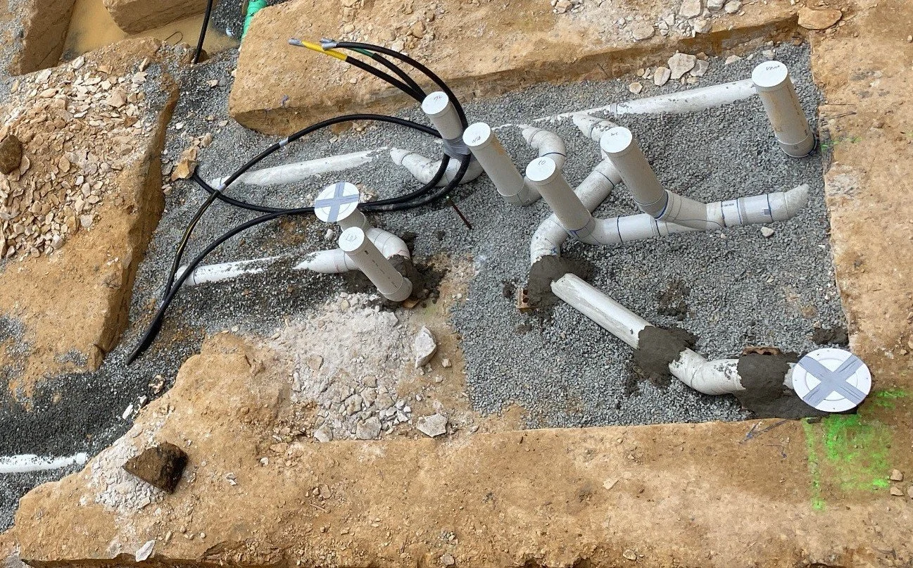 Underground plumbing pipes laid out in a trench with some black electrical wires and coverings over some pipe joints.