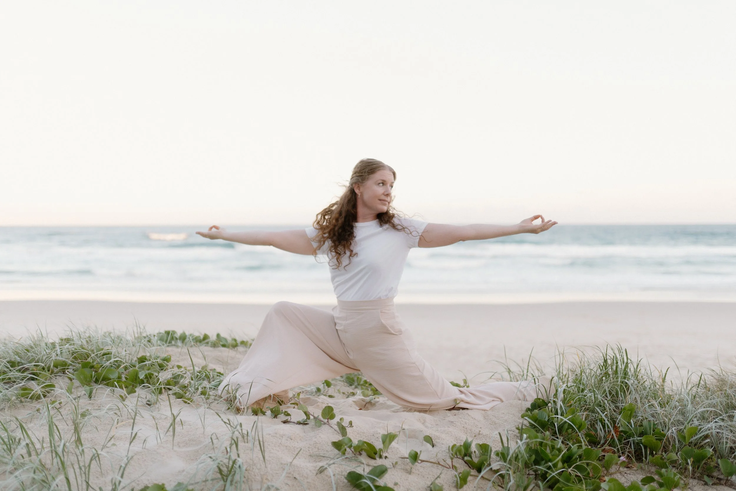 A woman with long curly hair practicing yoga in a lunge pose with arms extended on a sandy beach near the ocean, surrounded by green plants.