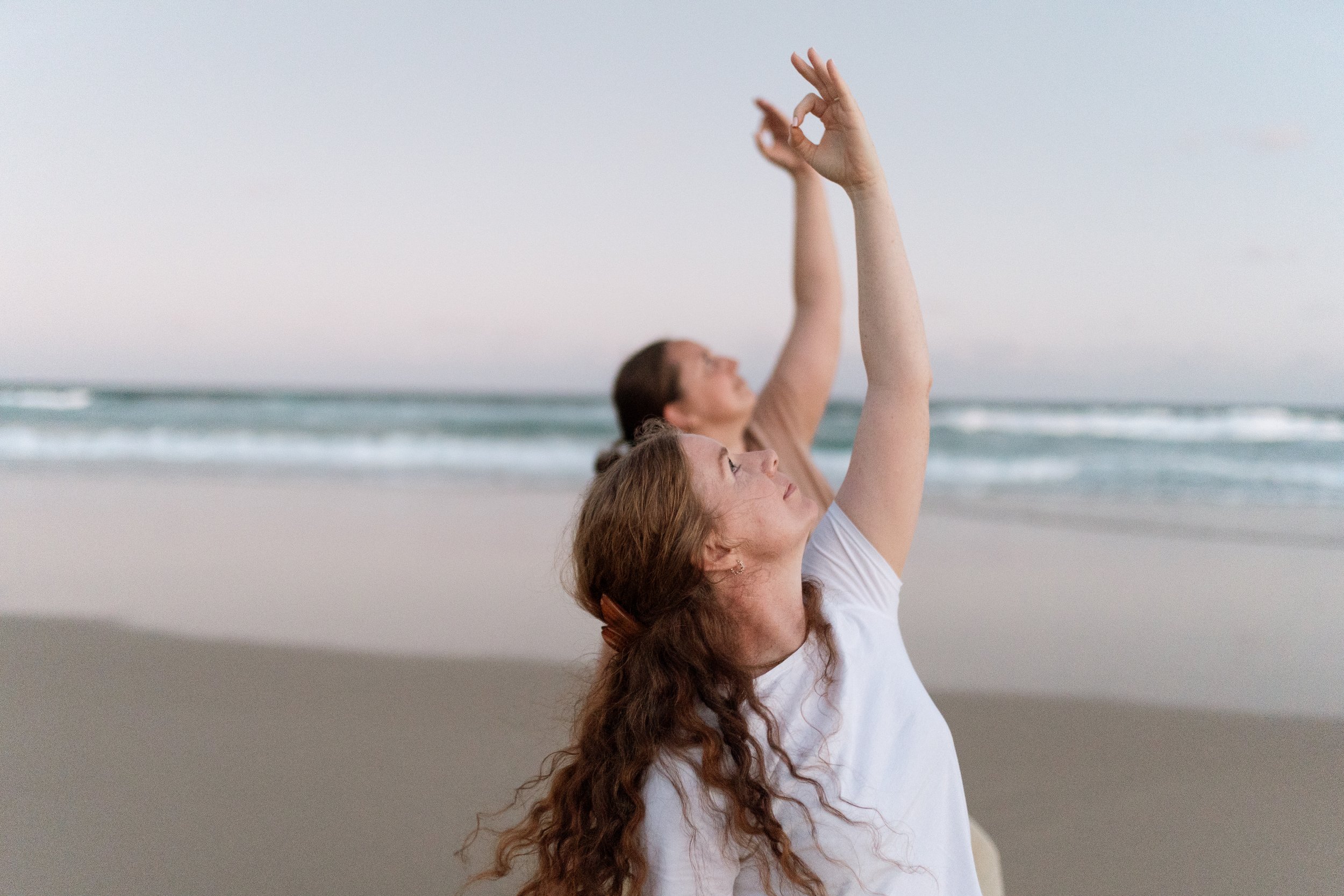 Two women practicing yoga on a beach, with hands raised and fingers forming an OK gesture, near the ocean with waves, during a sunset or sunrise.