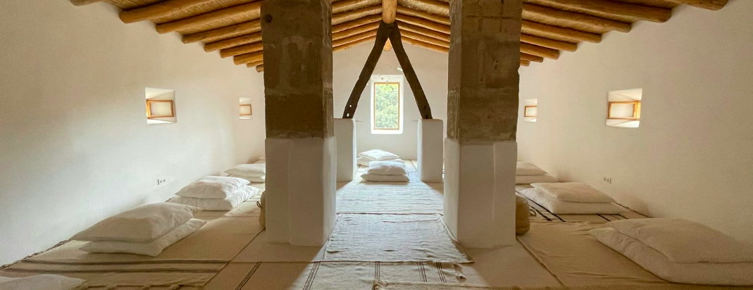 A minimalist, rustic attic with a pitched wooden ceiling, small windows, and pillows on the floor, arranged for sleeping along the walls.