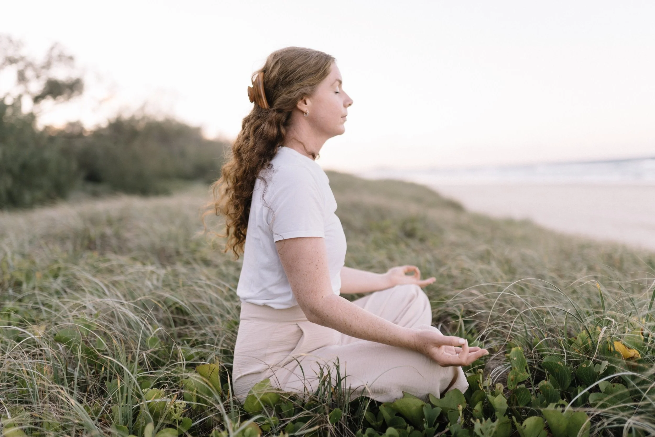 Woman with long curly hair meditating in a seated cross-legged position on a grassy beach, eyes closed, with a calm expression.