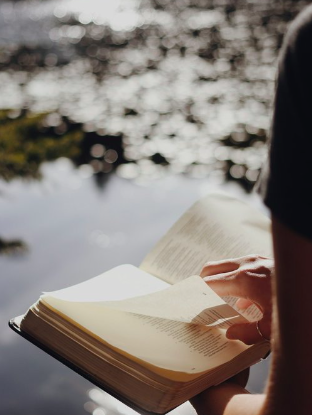 Person holding an open book outdoors near water.
