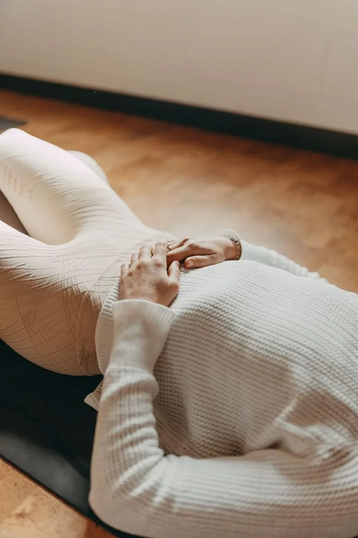 Person lying on a massage table with hands resting on their stomach, wearing a white sweater and light-colored pants, in a room with wooden flooring.