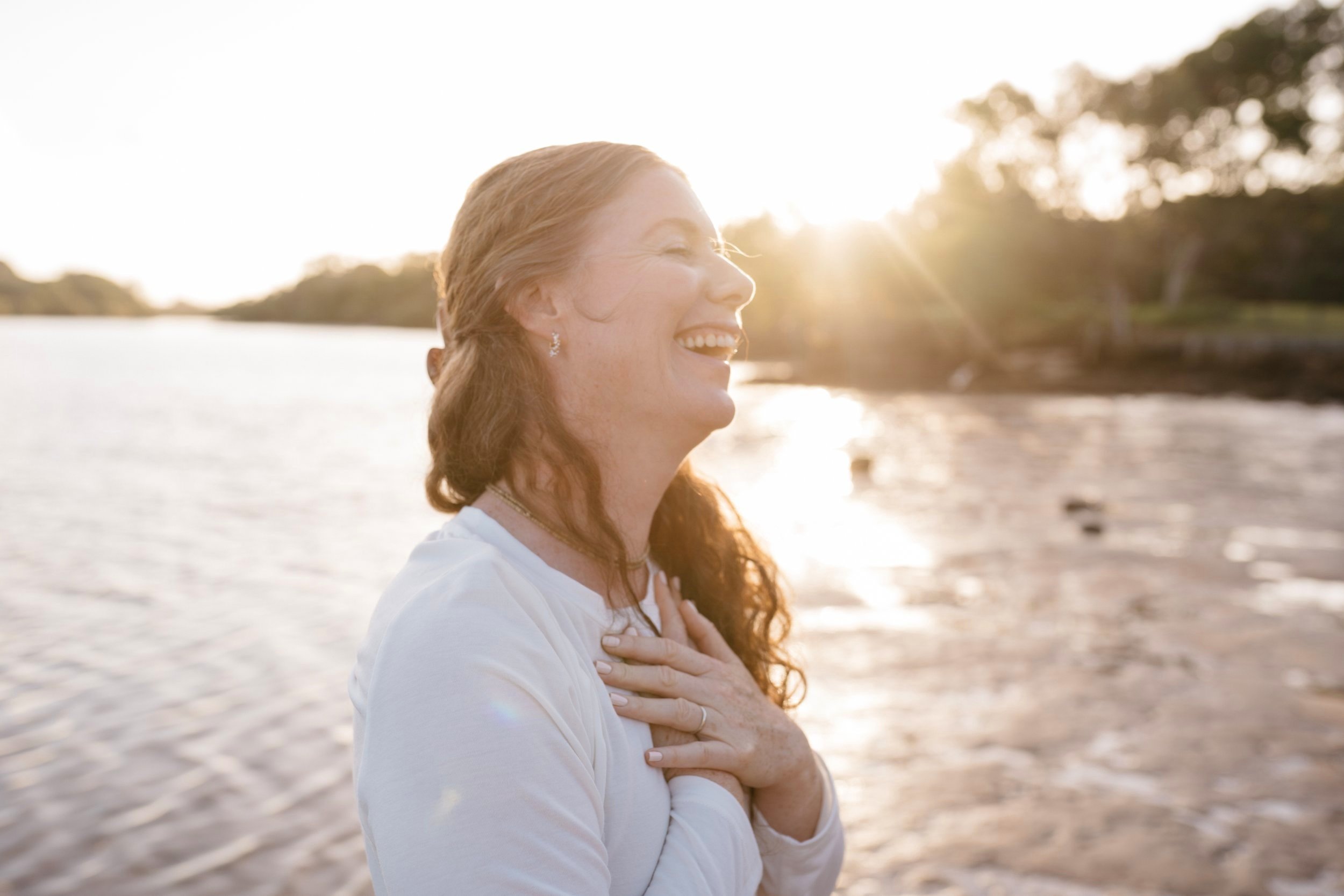 A woman with long, curly hair, smiling and holding her hands to her chest, standing on a beach at sunset.