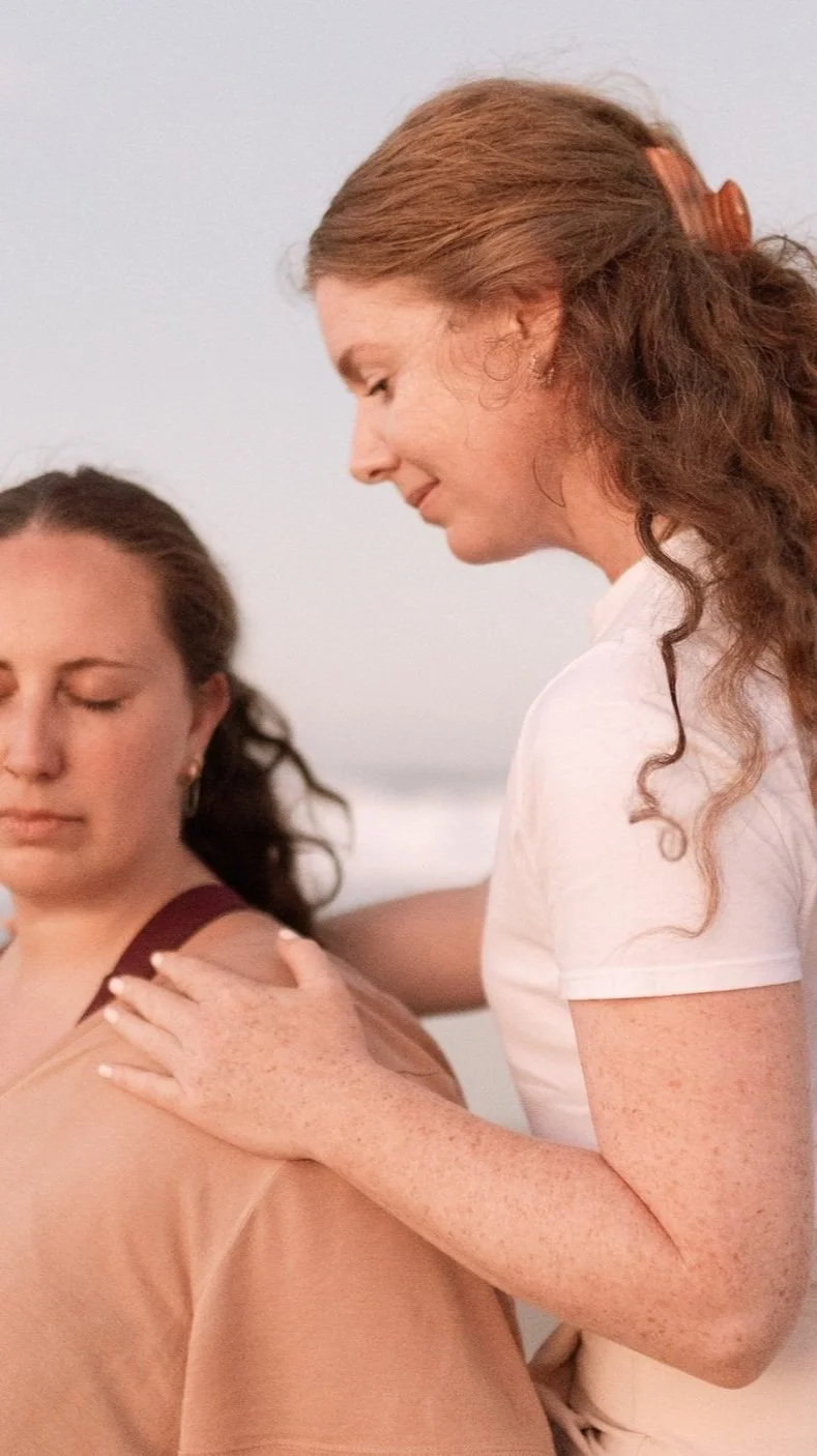 A woman with curly red hair and a white shirt gently places her hand on the shoulder of a woman with closed eyes, long dark hair, and a beige top, against a neutral background.