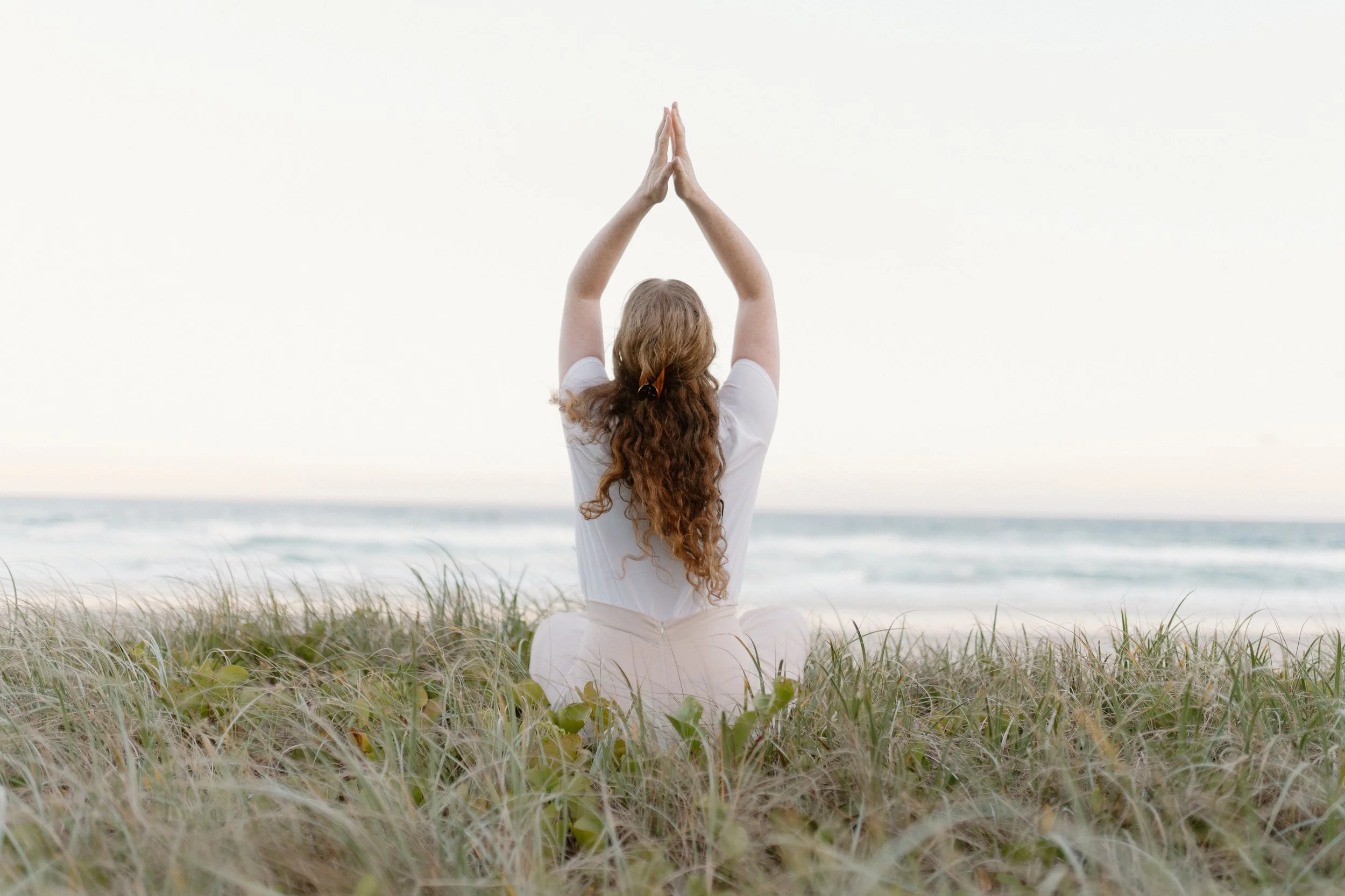 Woman with long, curly red hair practicing yoga on a grassy beach with ocean waves in the background, sitting cross-legged with arms raised and palms pressed together overhead.