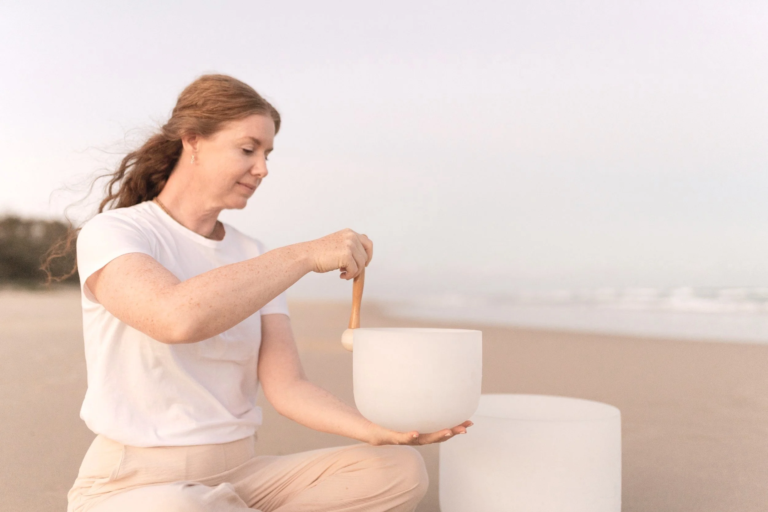 A woman with long red hair sitting on the beach, playing a white crystal singing bowl with a wooden mallet, during the daytime.