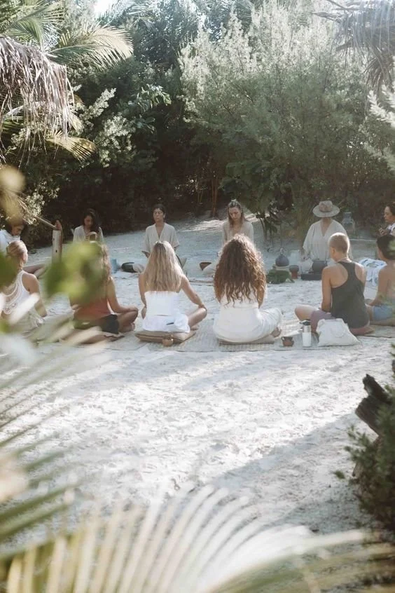 A group of people practicing meditation or yoga outdoors on a sandy surface, surrounded by lush green tropical plants, with some sitting cross-legged and one person leading the session, wearing a wide-brimmed hat.
