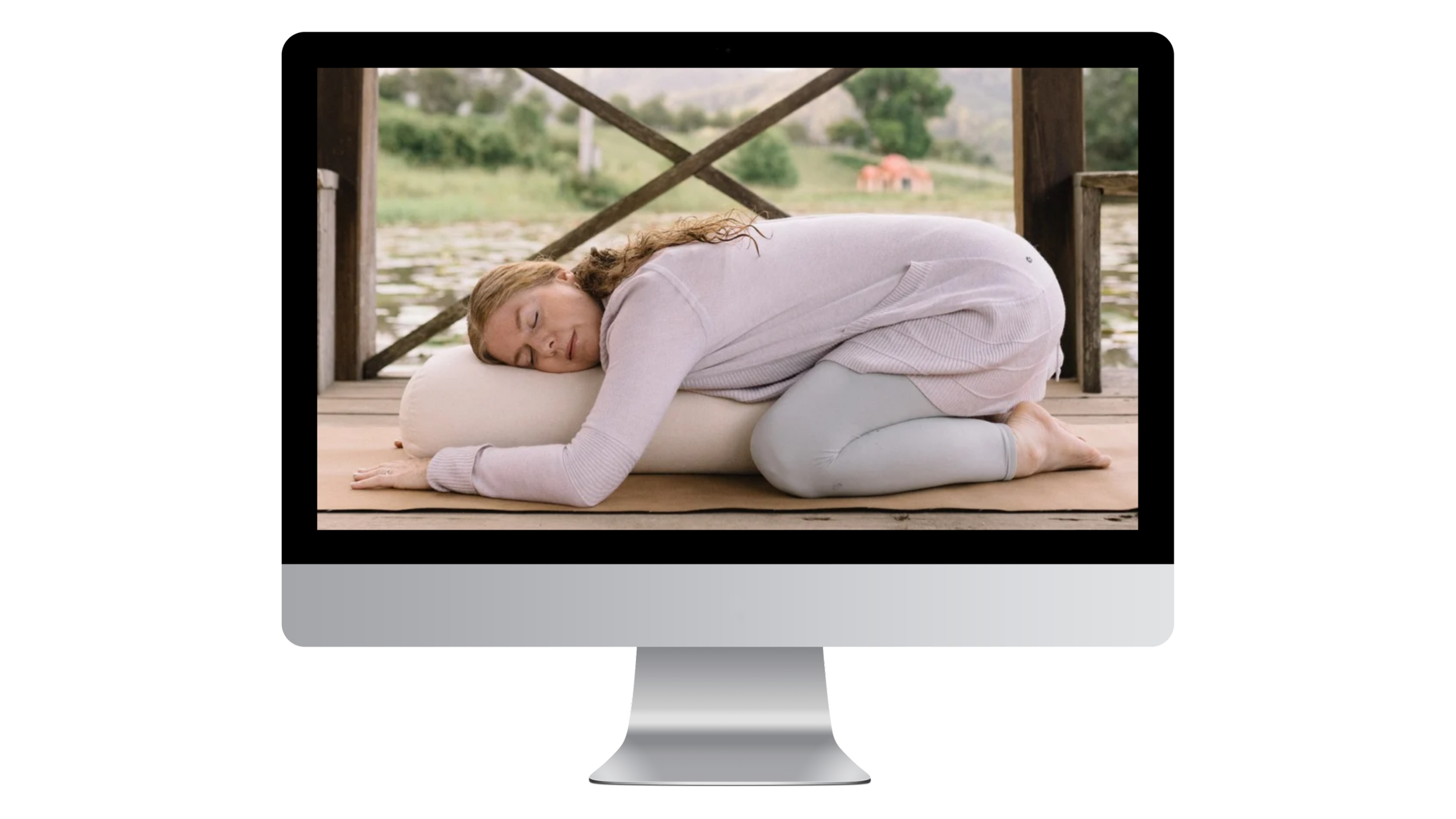 A woman practicing restorative yoga on a carpeted wooden dock by a lake, resting her head and arms on a rolled bolster with her eyes closed.