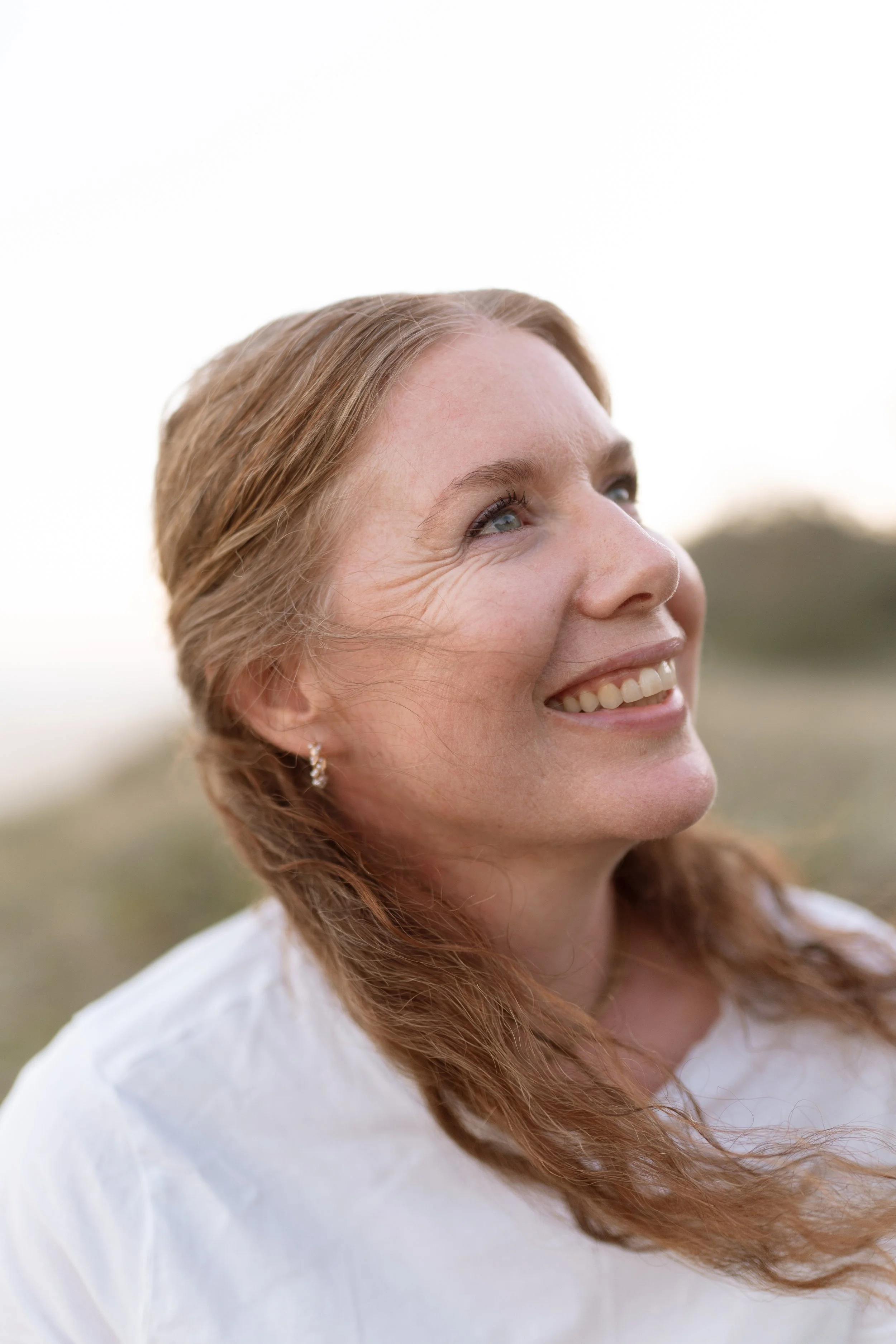 A smiling woman with red hair and earrings looking upward outdoors.