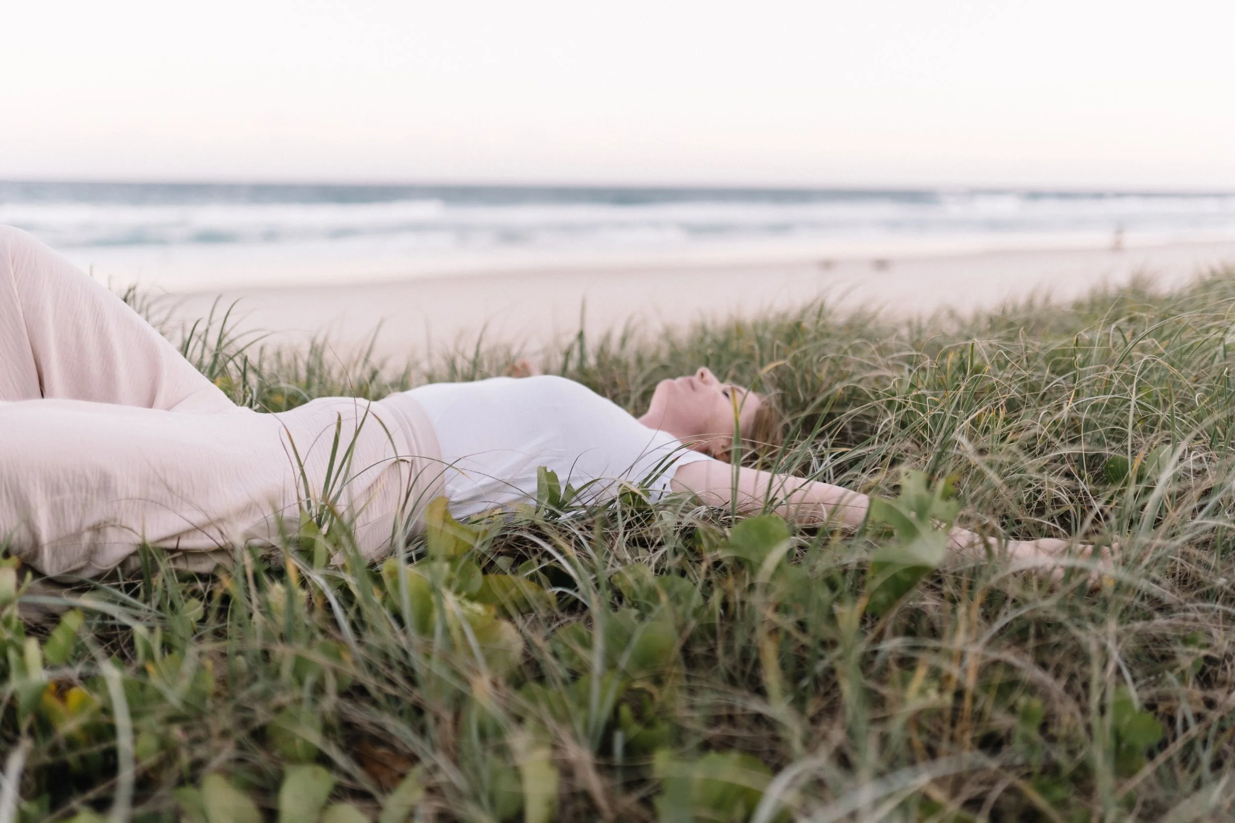 A woman lying on her back on a grassy area near the beach, with her eyes closed and arms stretched out, facing the sky, with the ocean and sand in the background.