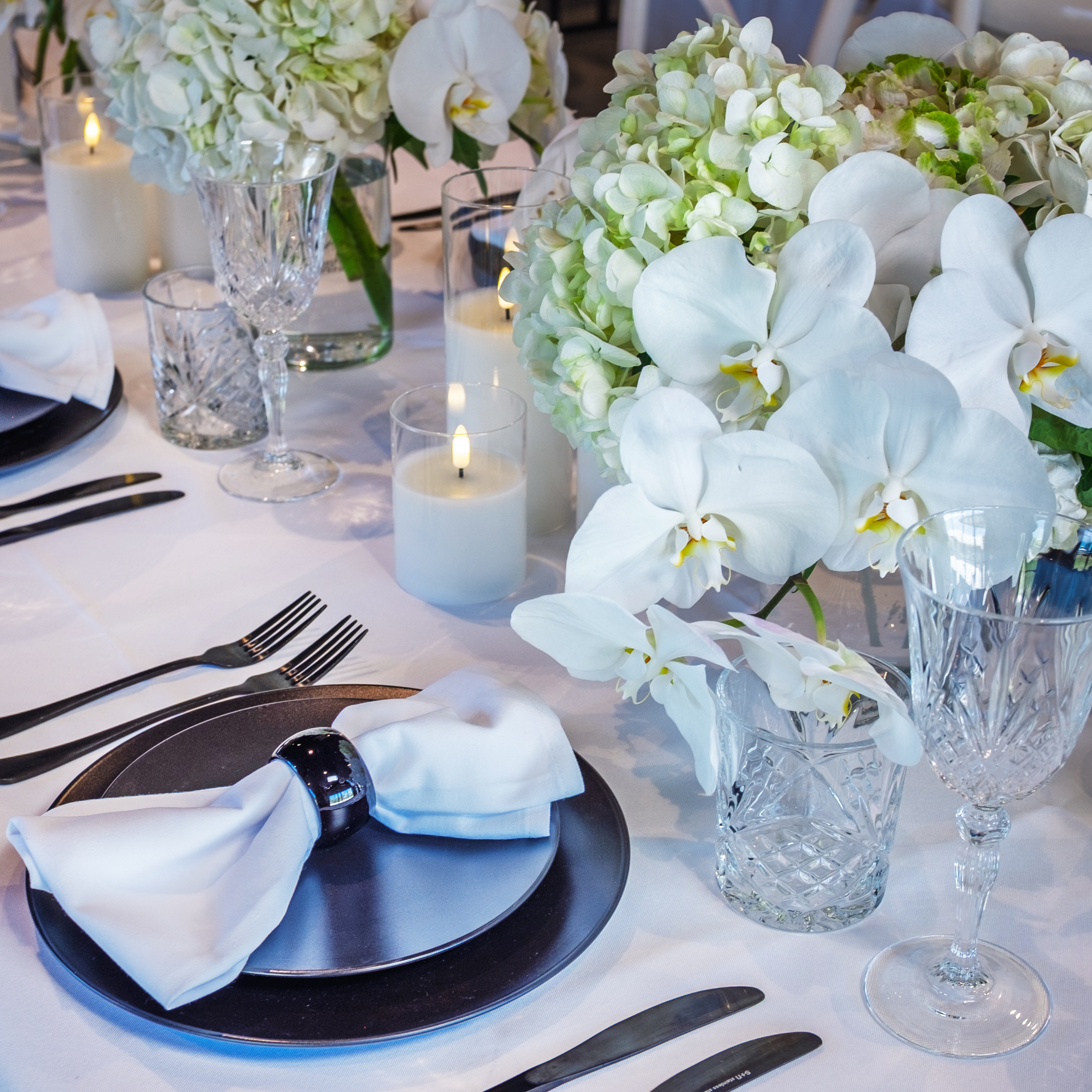 Elegant dining table setup with white floral centerpiece, glassware, candles, and black and white table setting.