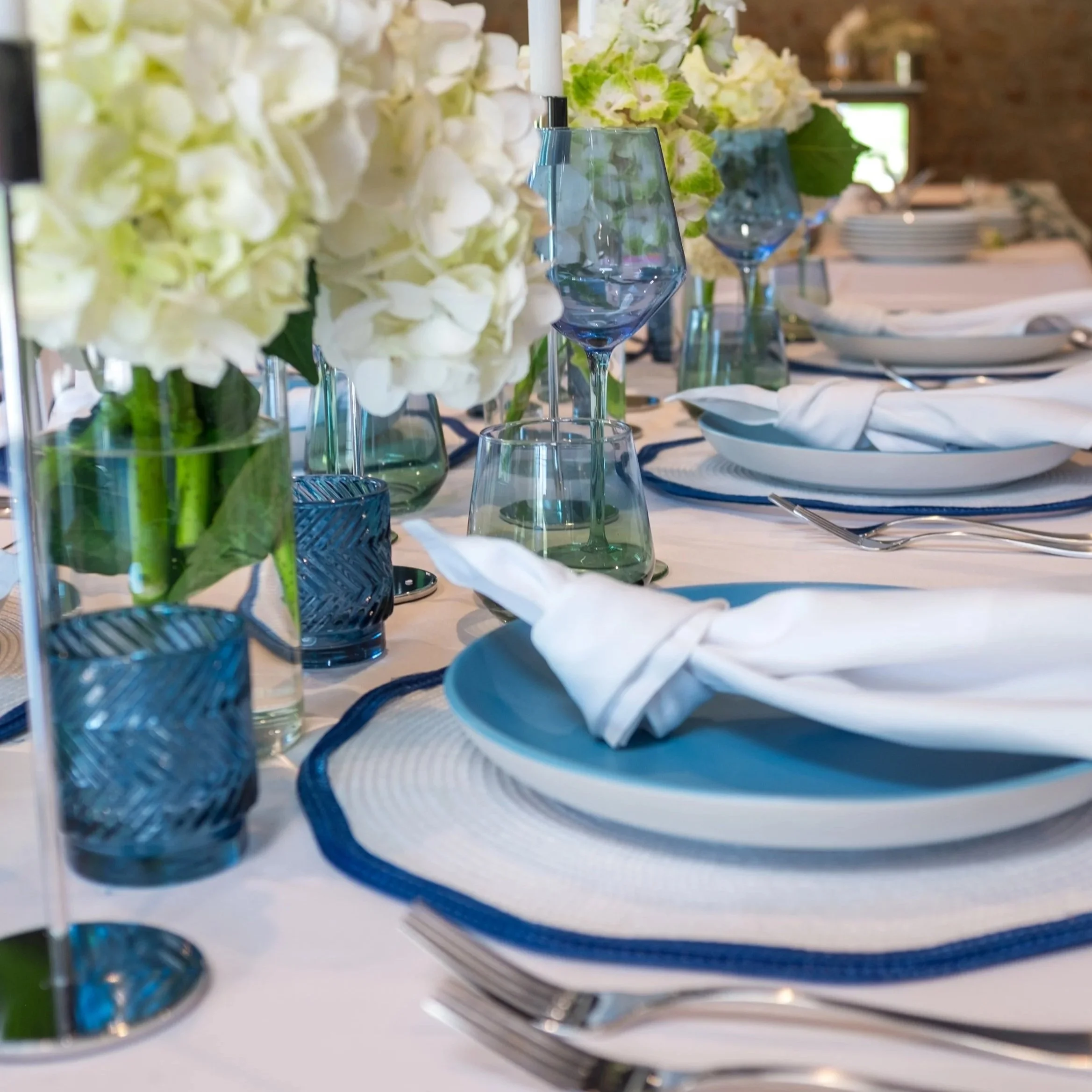 Elegant table setting with white and blue plates, white cloth napkins, glassware including blue-tinted glasses and wine glasses, floral centerpieces with white and green flowers, and a white tablecloth.
