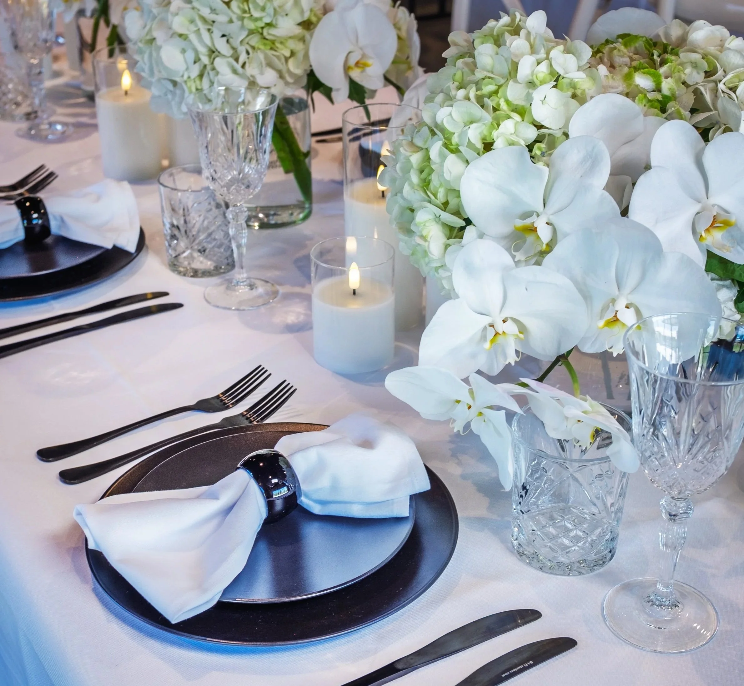 Elegant table setting with white napkin ringed with a black band, set on a black plate, surrounded by black utensils, crystal glasses, white candles, and a large floral centerpiece of white orchids and hydrangeas.