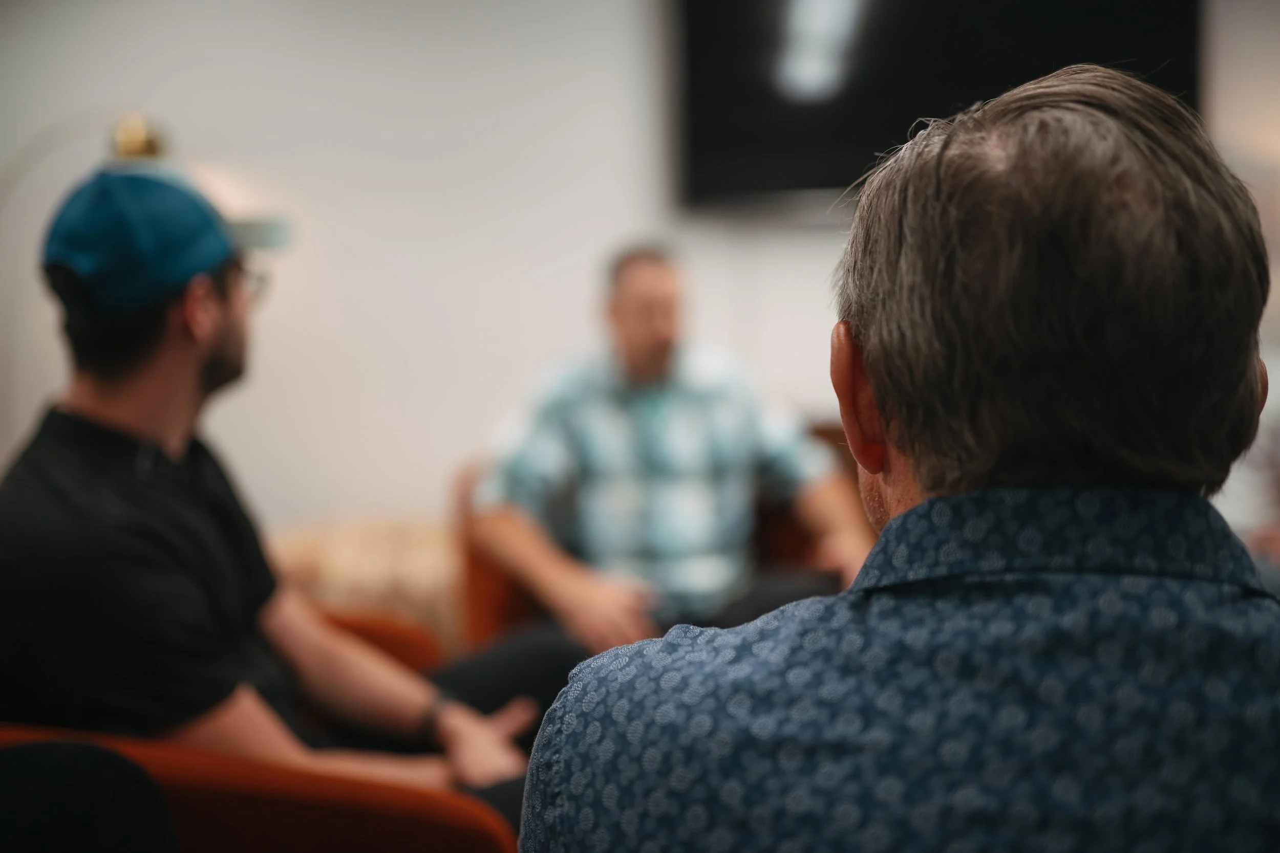 Three men in a meeting room, one in focus with a patterned shirt, two others blurred in the background, engaging in a discussion.