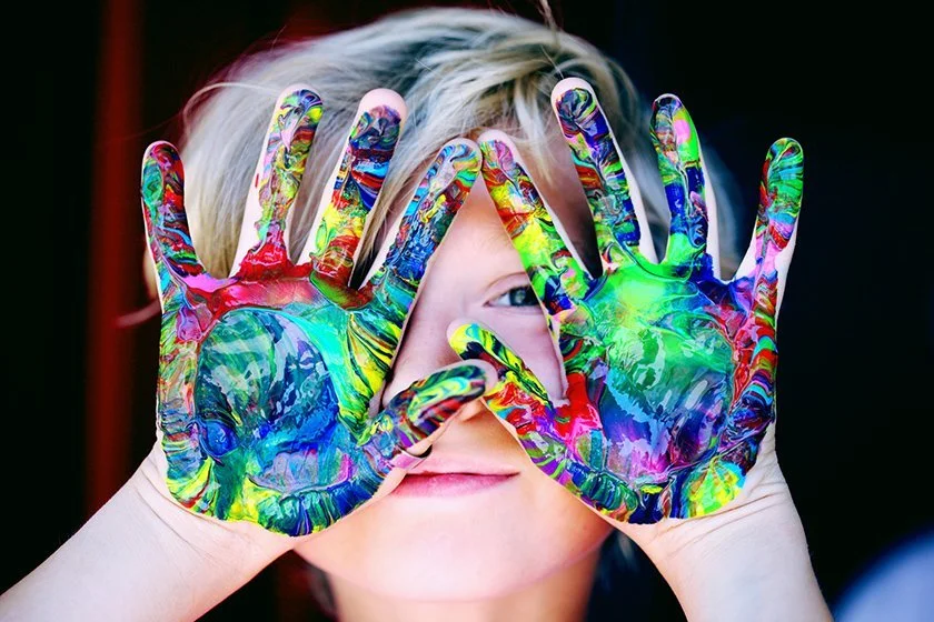 Child with painted hands holding them up in front of face, smiling.