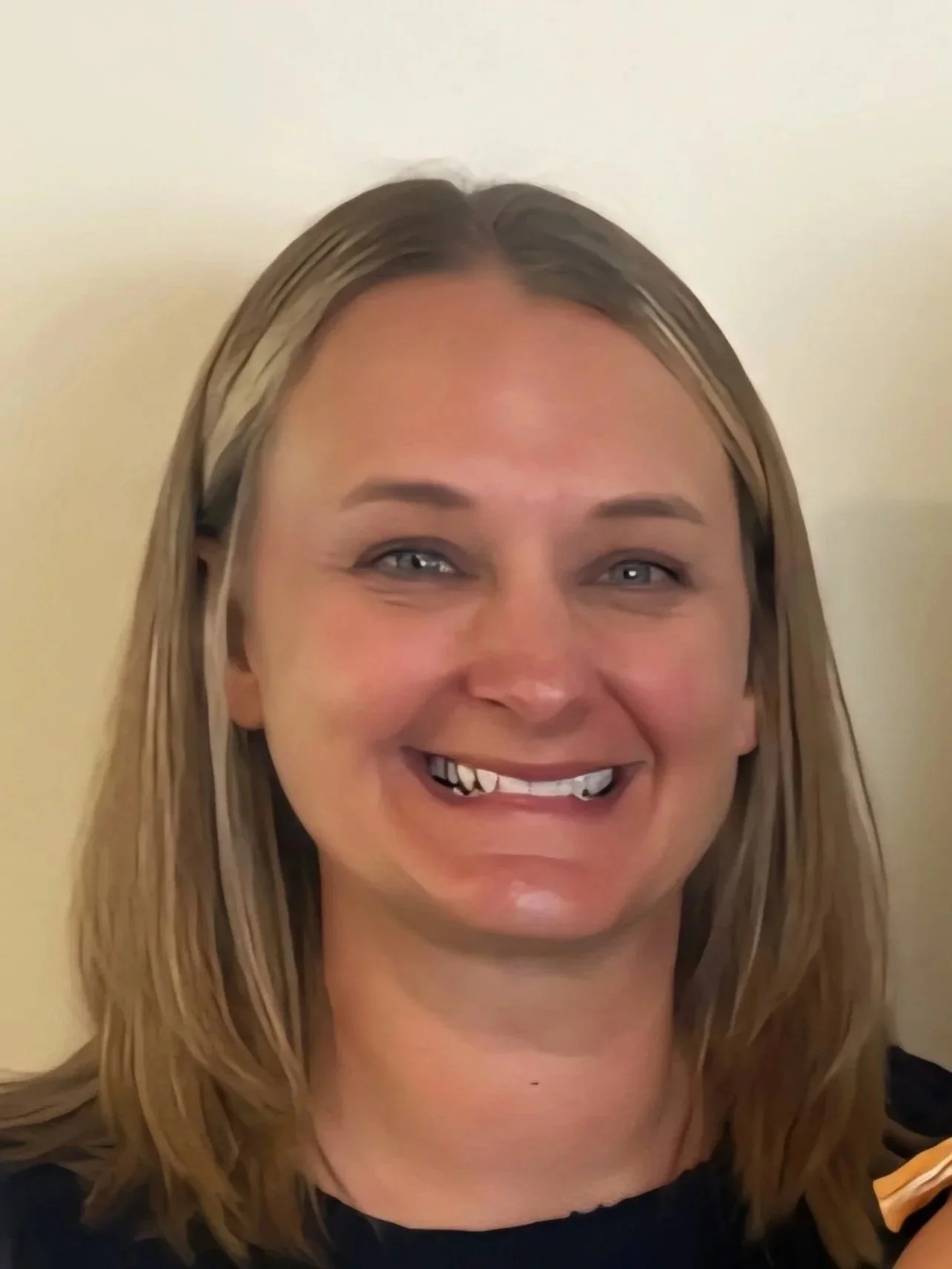 A woman smiling, with some teeth missing, and blonde hair, standing against a plain wall.