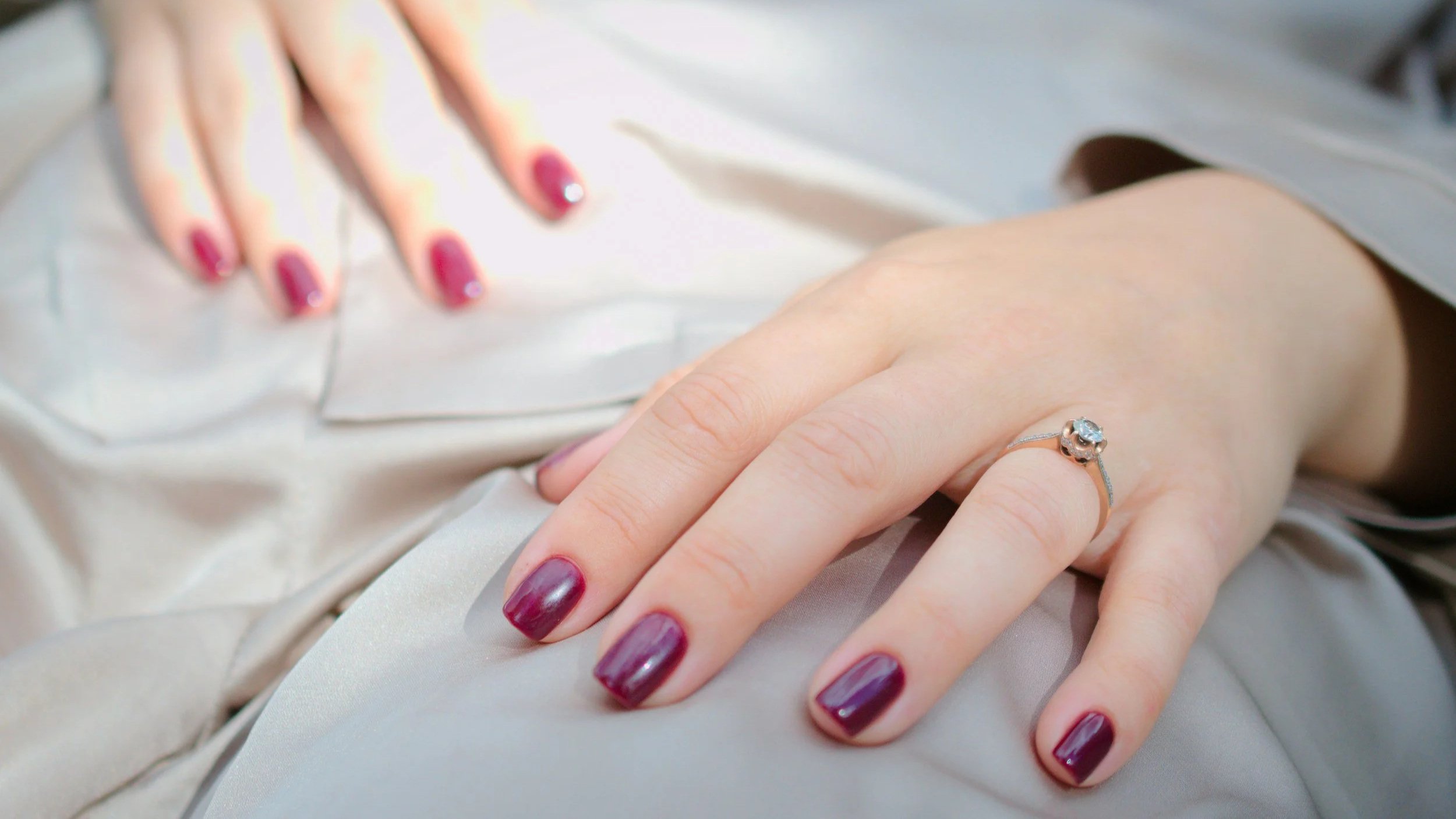 Close-up of a woman's hand with maroon nail polish, resting on a beige satin fabric, wearing a silver ring with a clear gemstone.