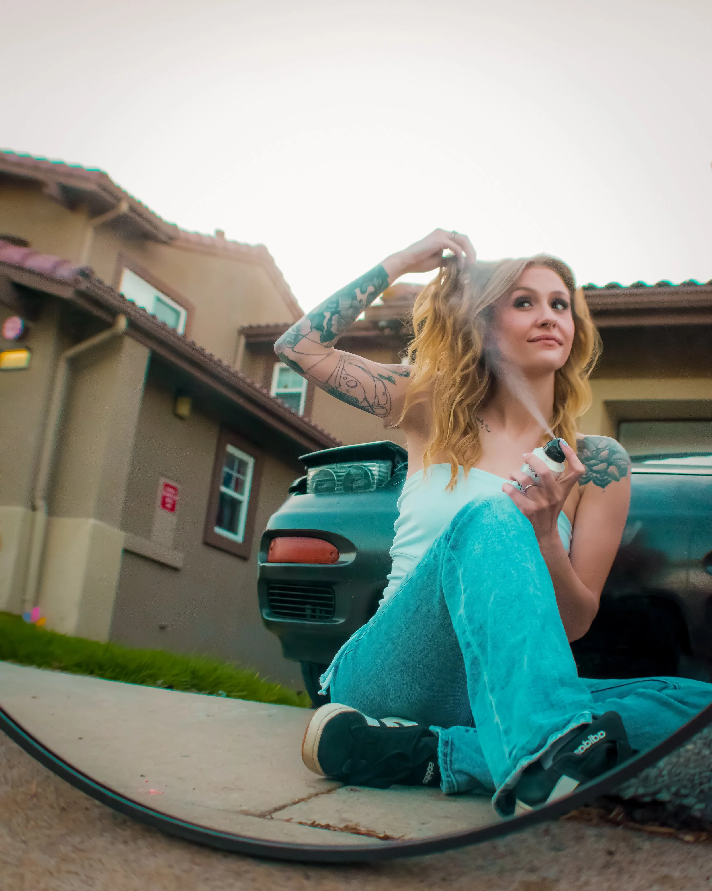 Young woman with tattoos in a white tank top sitting on the sidewalk, spraying perfume, with a house and parked car in the background, reflected in a mirror.