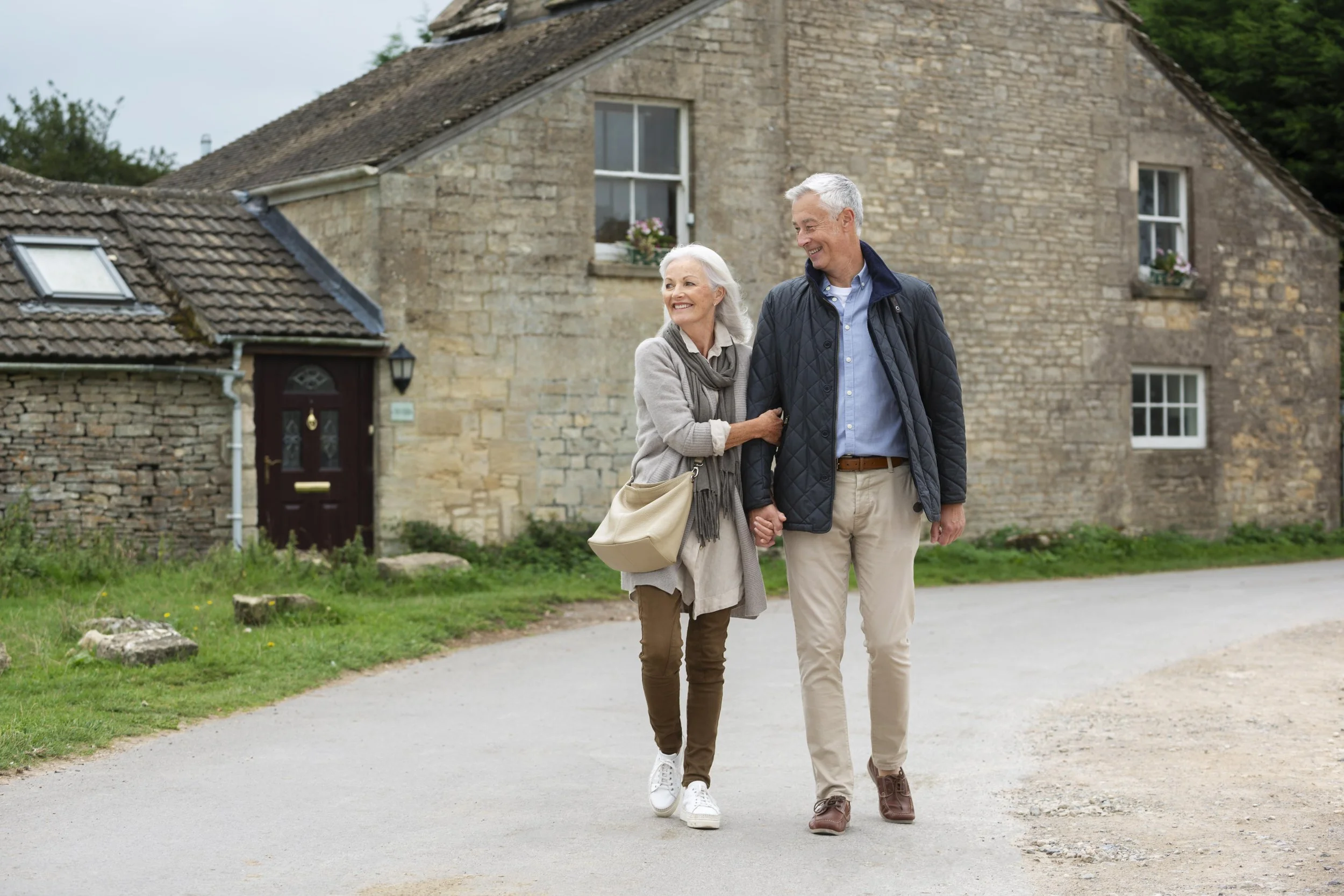 An elderly couple walking hand in hand along a country road, smiling and looking at each other, with a stone cottage in the background.