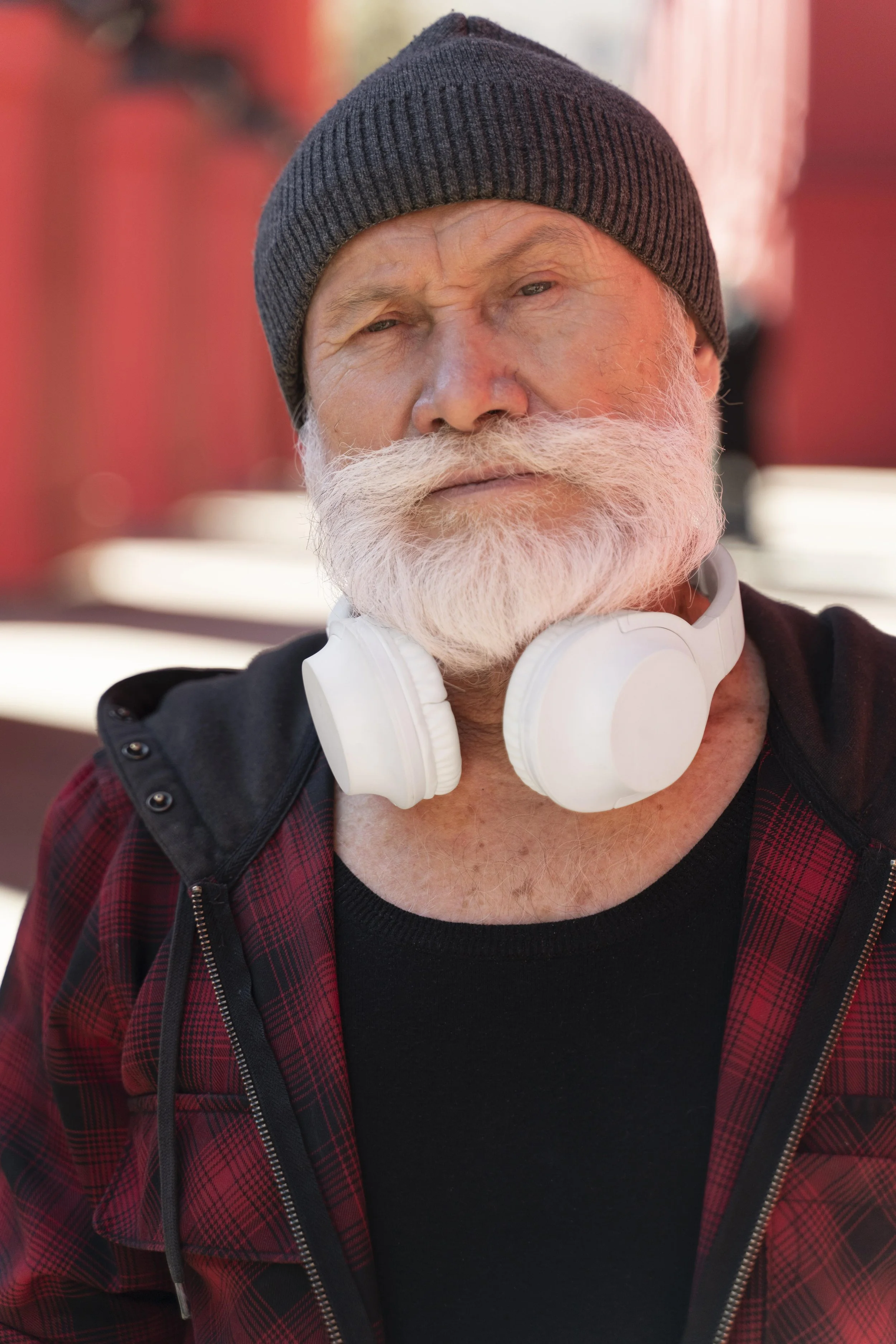 A young man with dark hair and light skin, wearing round sunglasses, a brown button-up shirt over a white T-shirt, standing outdoors in front of a brick wall with a dark window in the background.