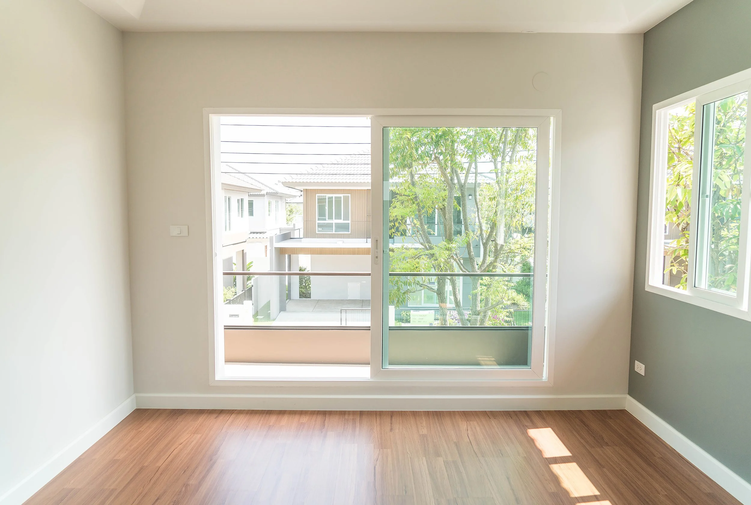 Empty room with large sliding glass door and window, hardwood floors, and views of trees and neighboring houses.