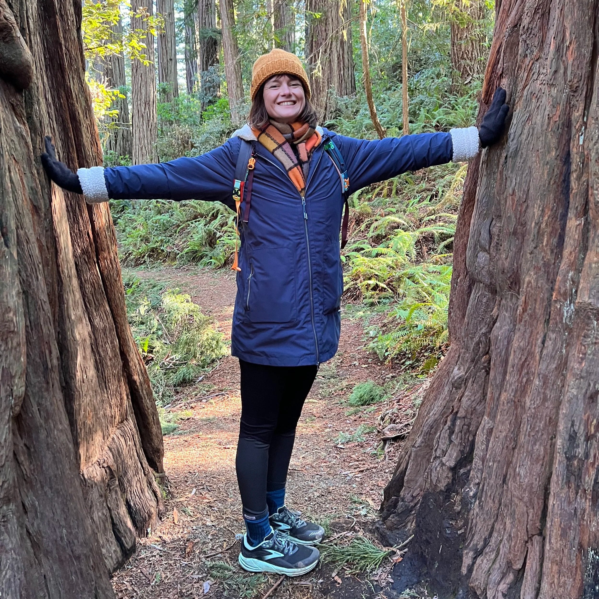 A woman standing on a forest trail, smiling with arms outstretched, hugging two large tree trunks.