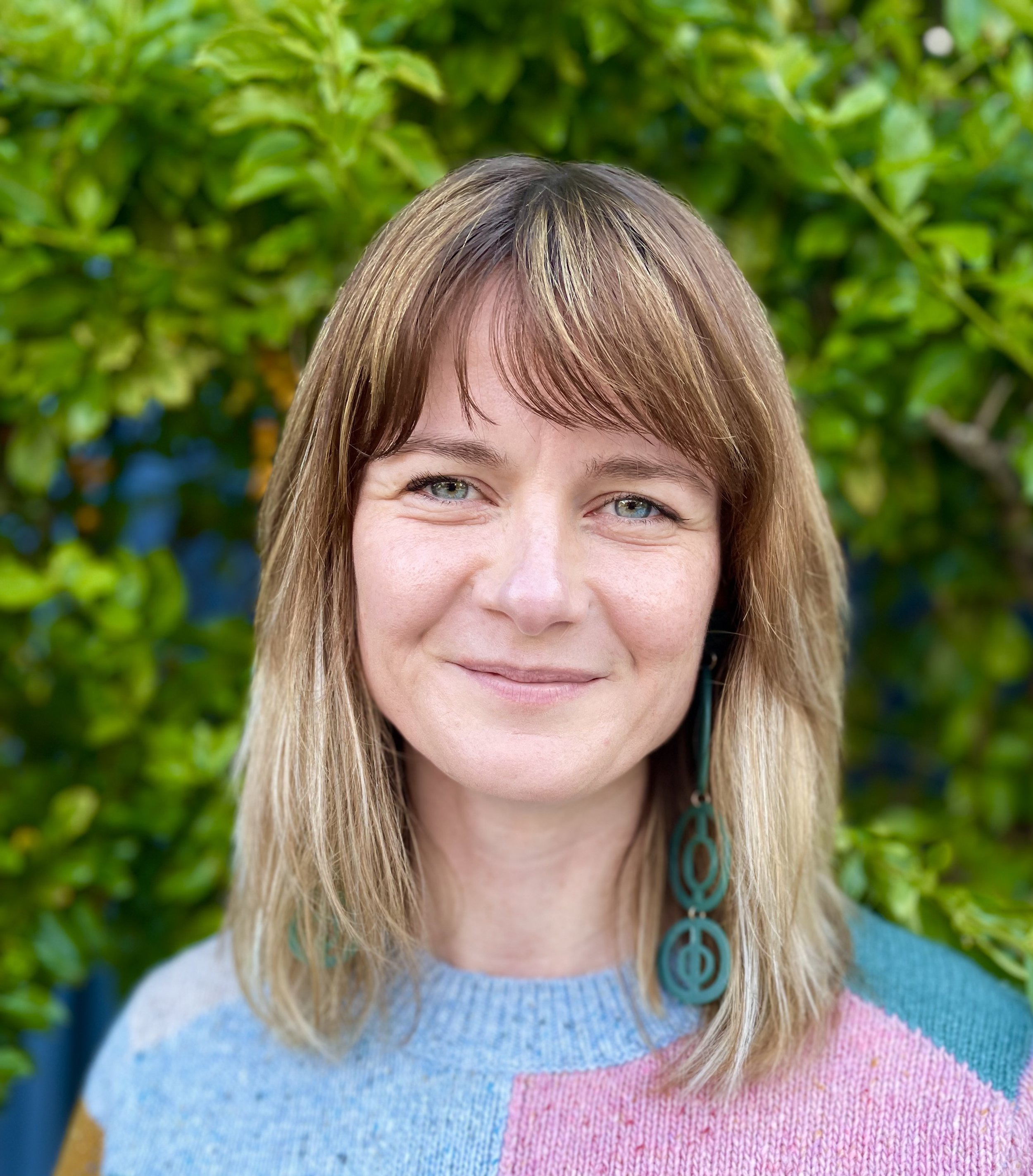 Close-up of a woman with shoulder-length blonde hair and blue eyes, smiling outdoors with green foliage in the background, wearing a multicolored sweater and large teal earrings.