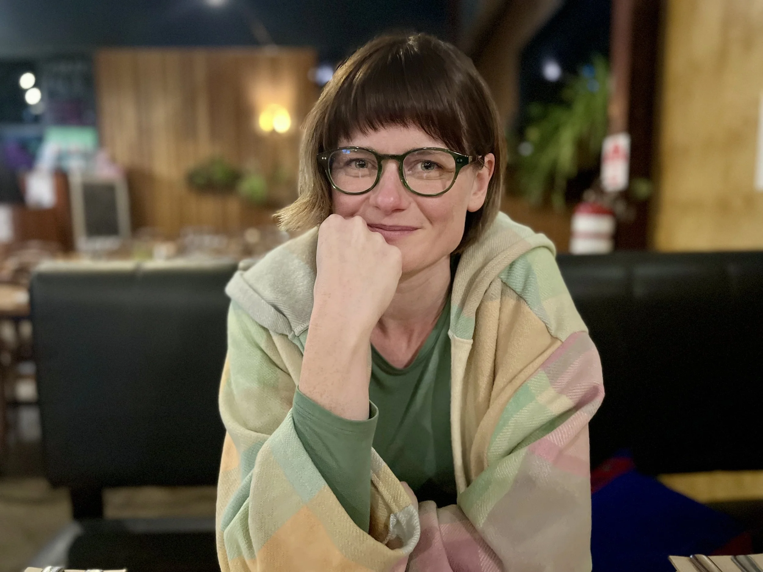 A woman with short brown hair and glasses sitting at a restaurant, resting her chin on her hand, smiling at the camera. She is wearing a green shirt and a multicolored blanket or shawl.