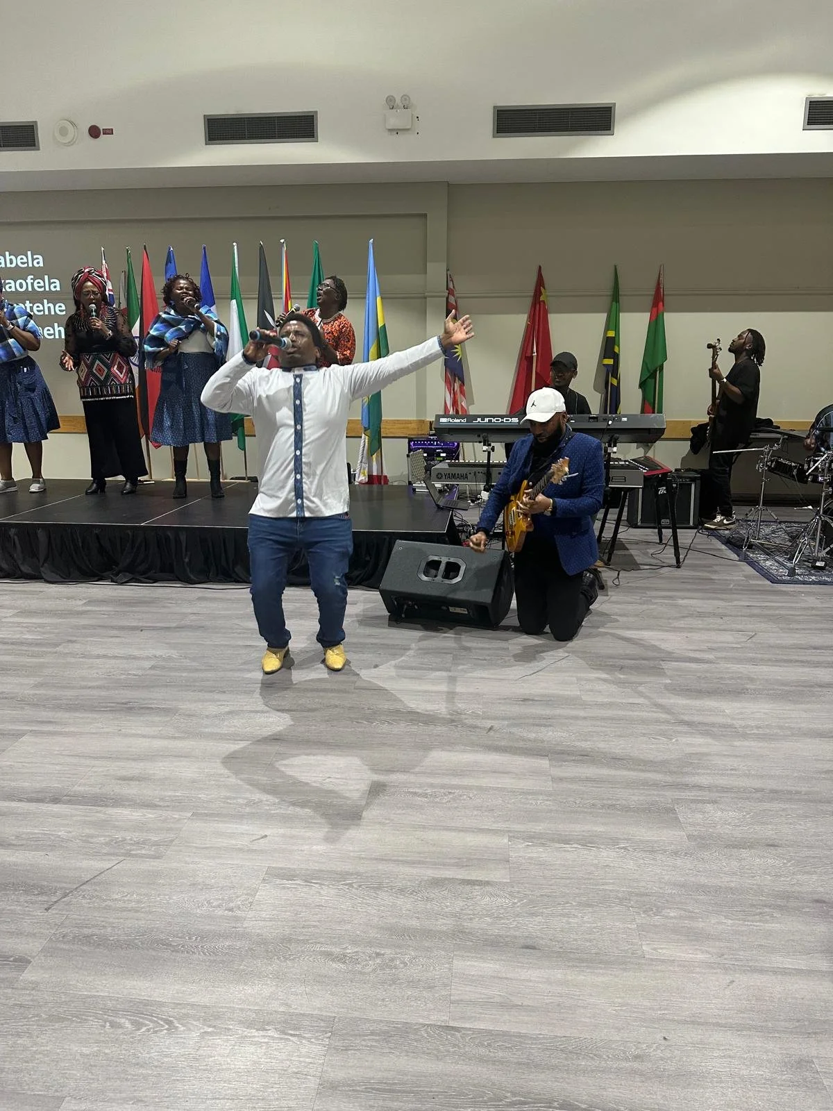 A gospel music performance with a man singing into a microphone, several women singing on stage, and musicians playing instruments, with multiple international flags in the background.