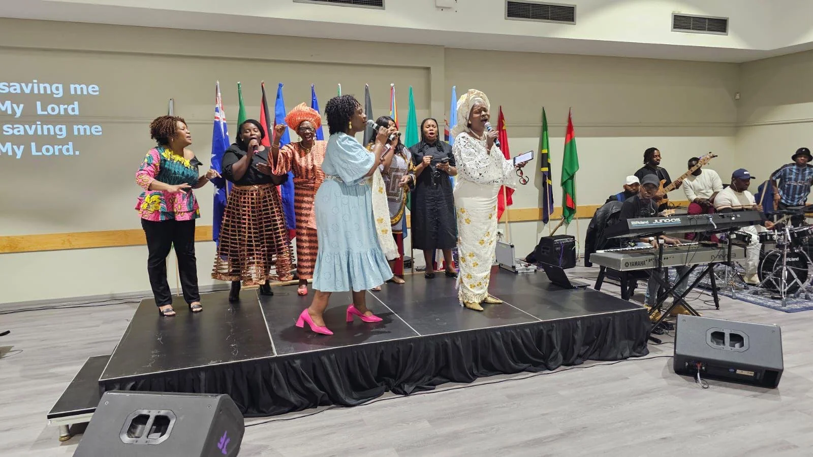 Group of women singing on a stage with musical instruments and flags in the background.