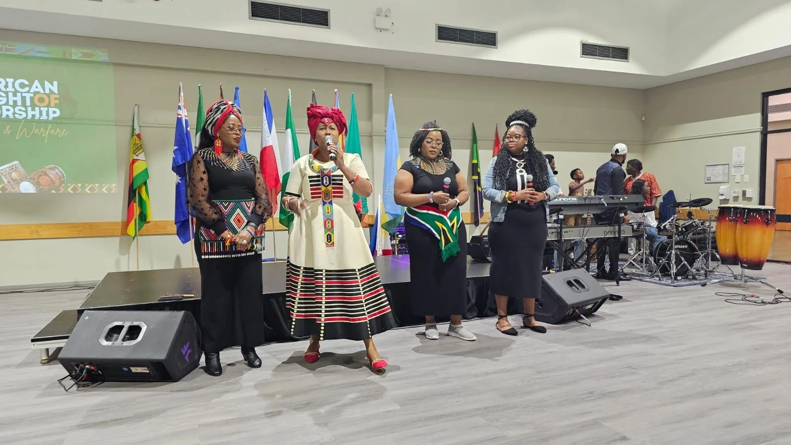 Four women dressed in vibrant cultural attire stand on stage during a presentation or event, with several national flags behind them and musical instruments to the right.
