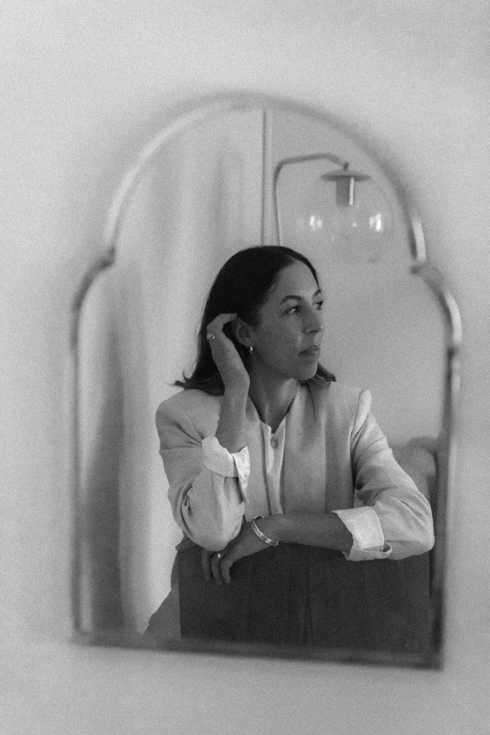 A woman is looking into a mirror, adjusting her earring, with a contemplative expression, in a black and white photo.