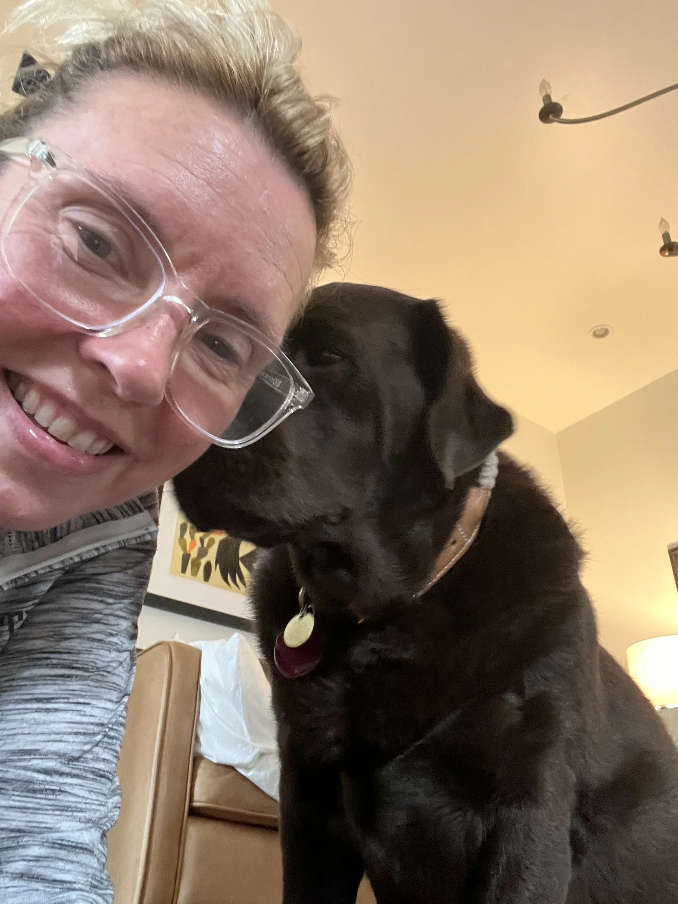 A woman with glasses smiling and a black dog with a collar close together indoors.