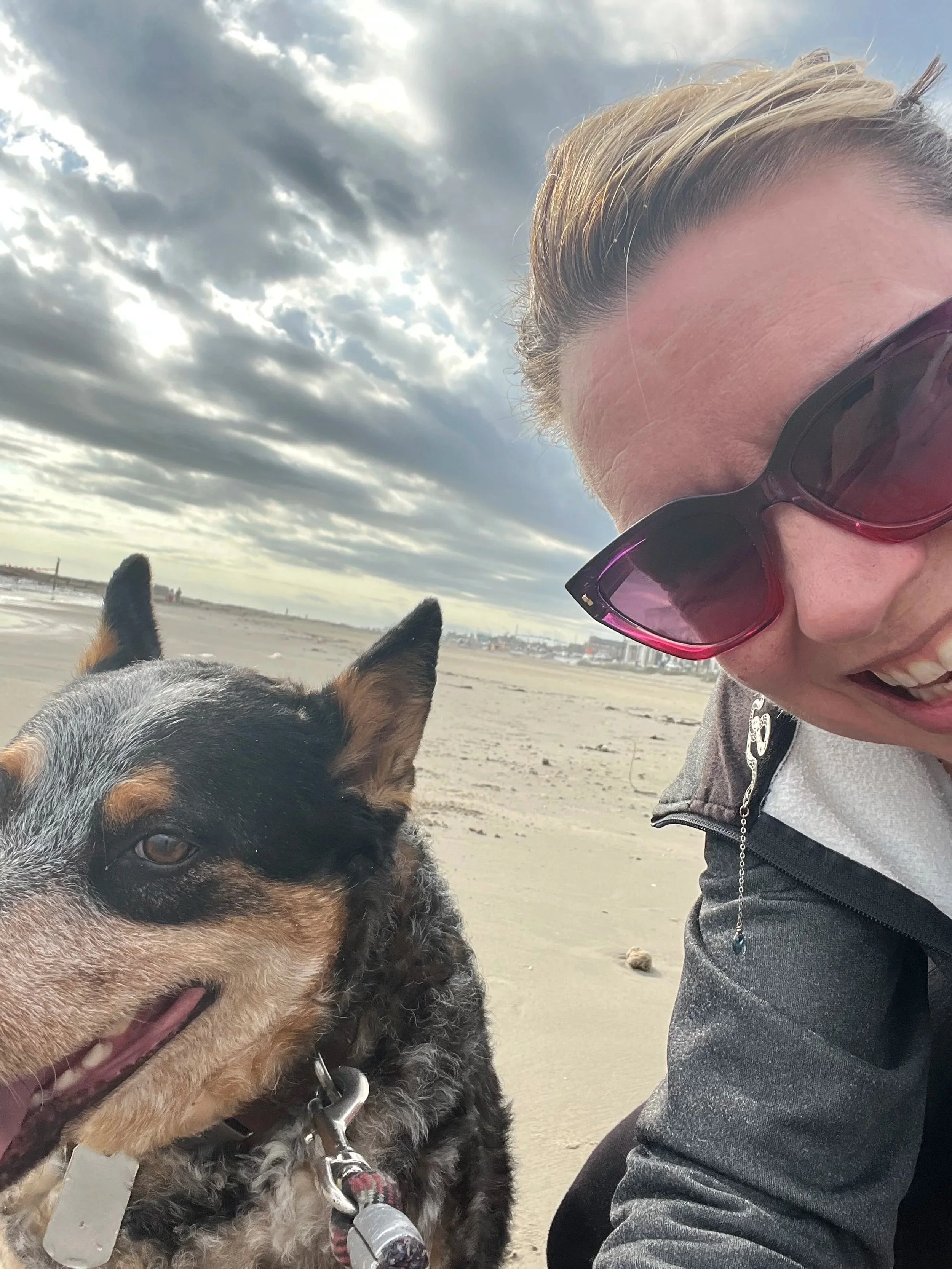 A woman wearing sunglasses taking a selfie on a beach with a dog, both smiling, overcast sky with clouds.