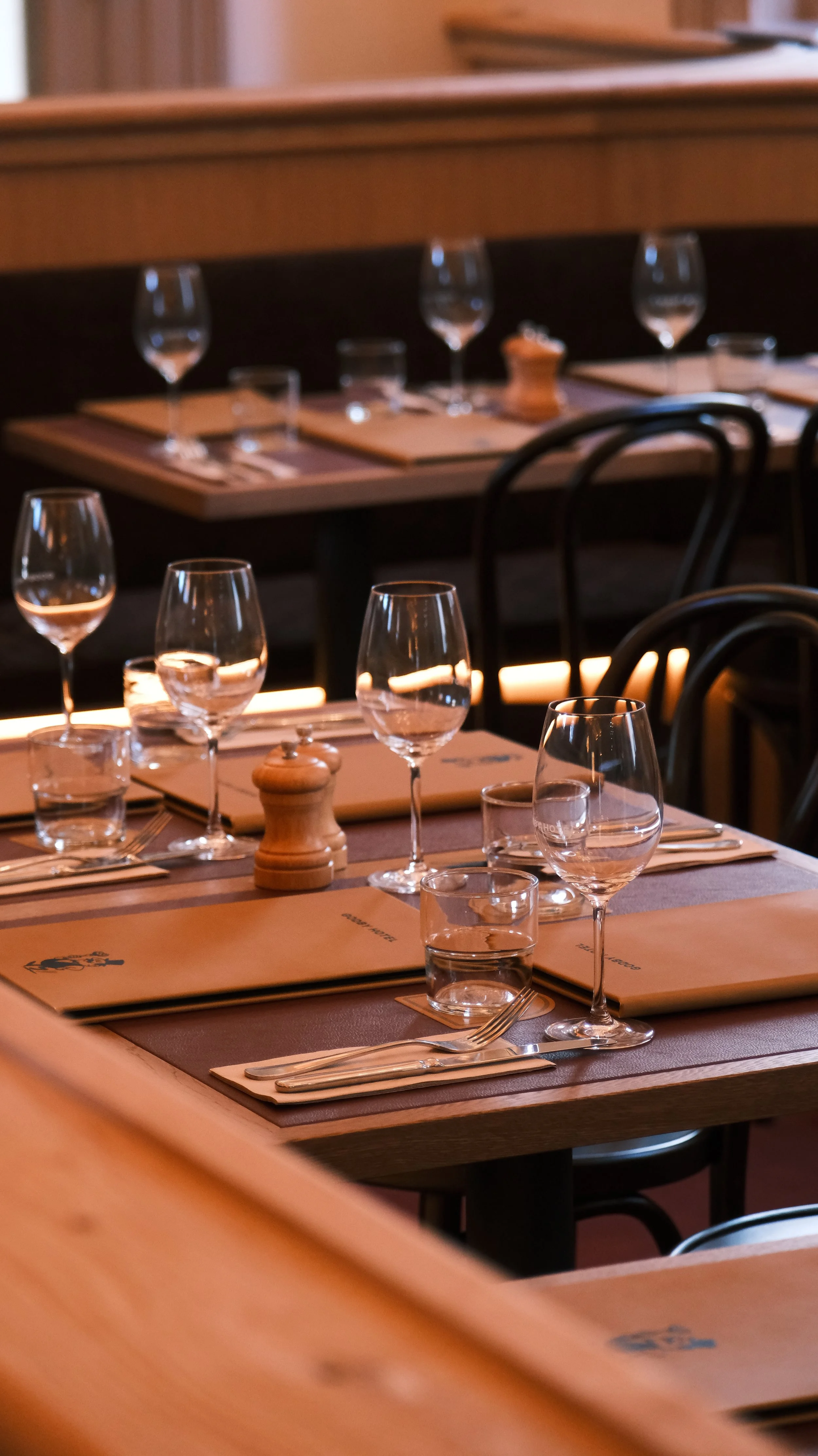 Restaurant table setting with wine glasses, water glasses, a pepper grinder, menus, cutlery, and napkins, in a warmly lit dining area.
