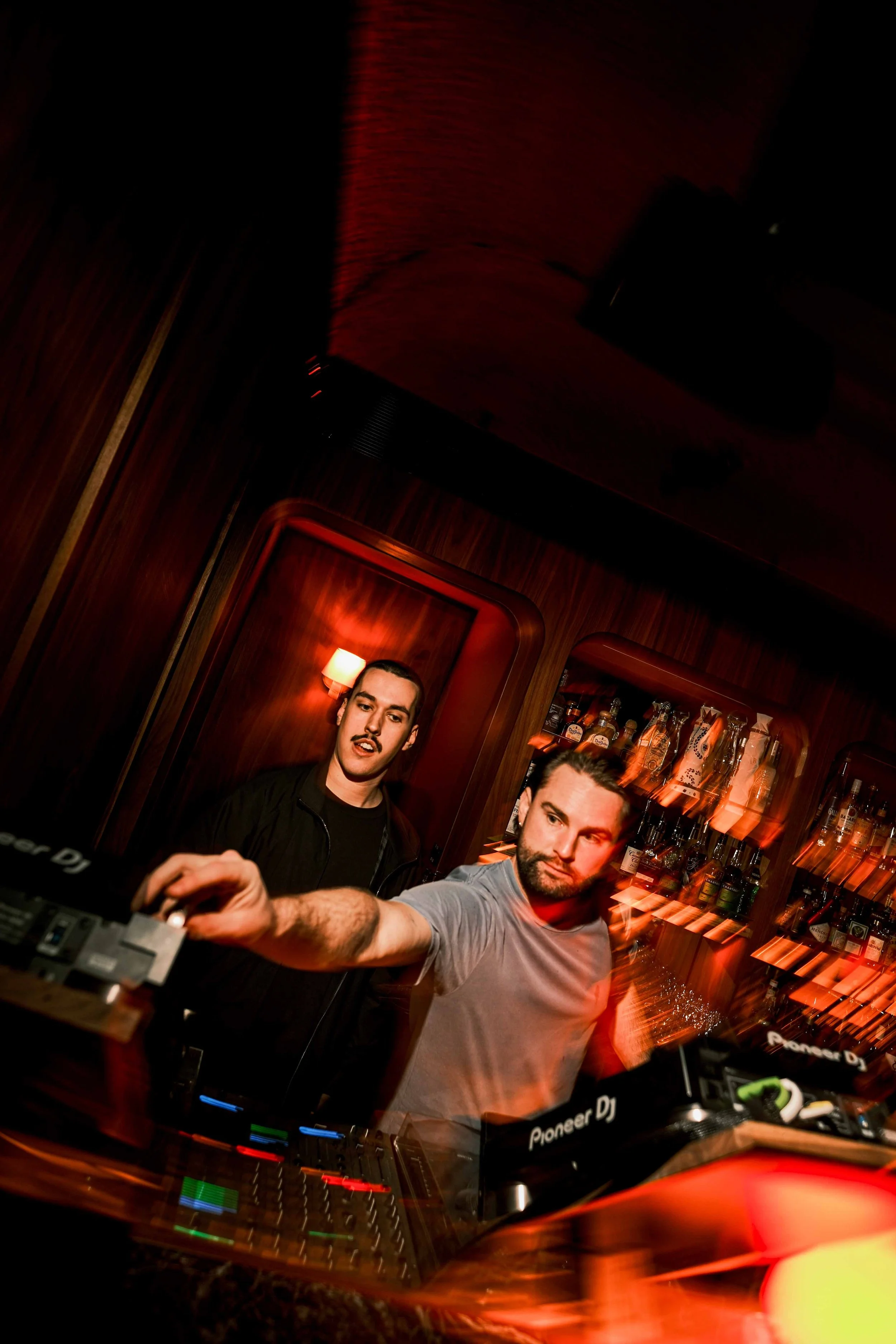 A DJ with tattoos on his arms wearing a black shirt, with headphones, is mixing music behind a DJ booth in a bar or club. There are bottles on shelves in the background and a woman with hoop earrings is visible on the right.