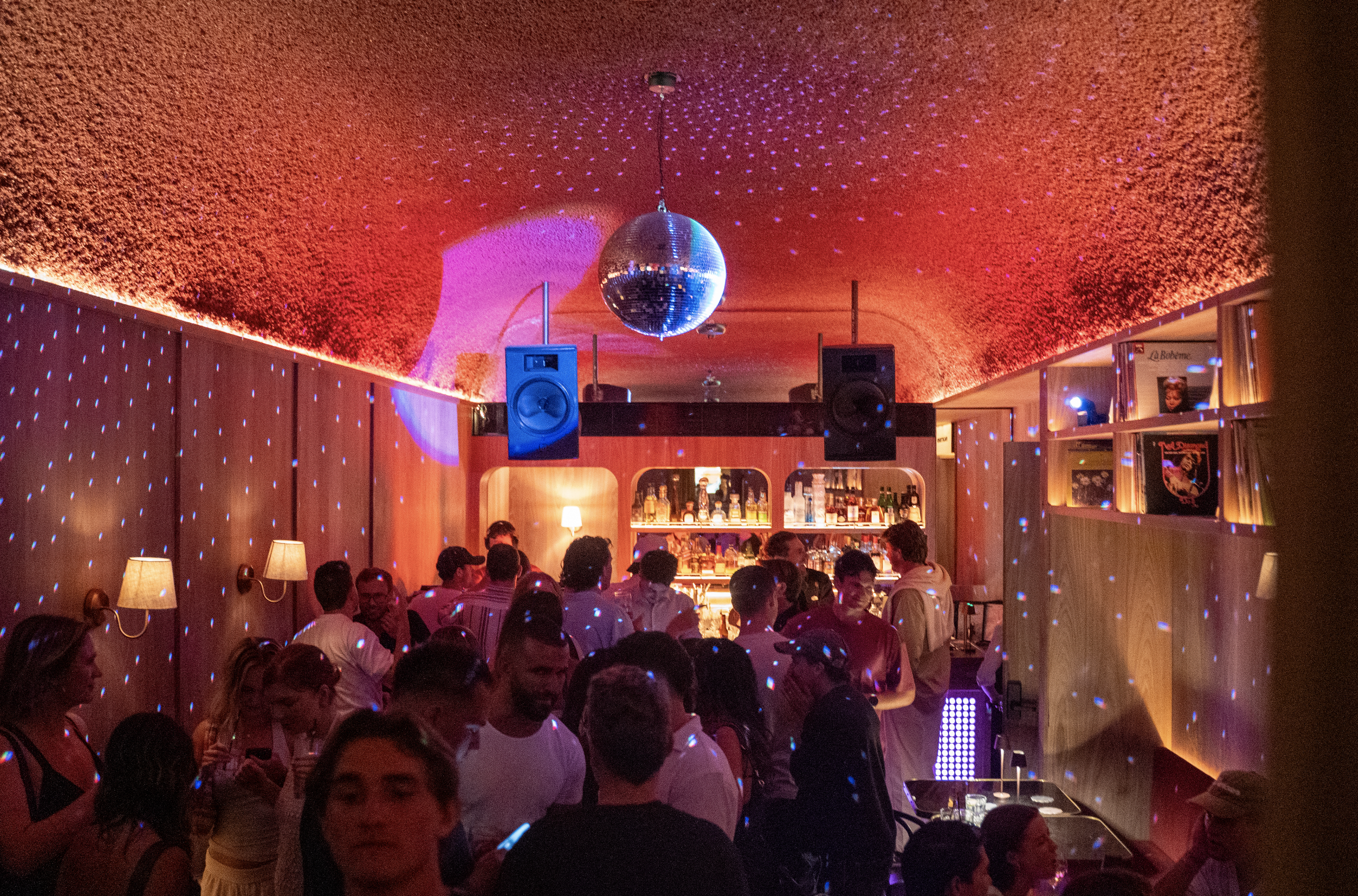 People sitting and talking at a bar or restaurant with wooden walls, shelves of bottles in the background, and a disco ball hanging from the ceiling.