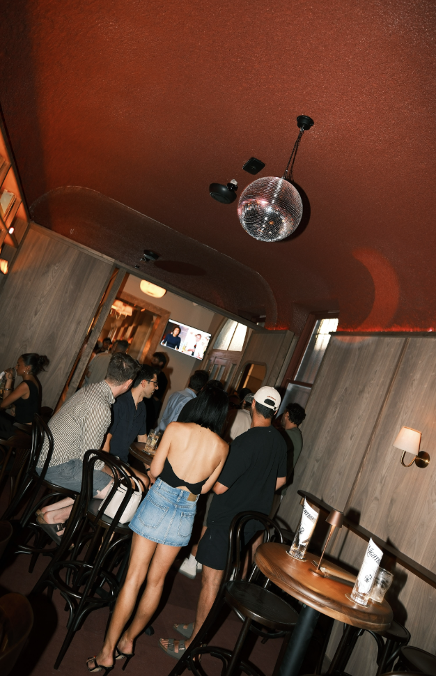 People gathered inside a bar or nightclub, watching a television screen, with a woman in a black top and denim skirt in the foreground.