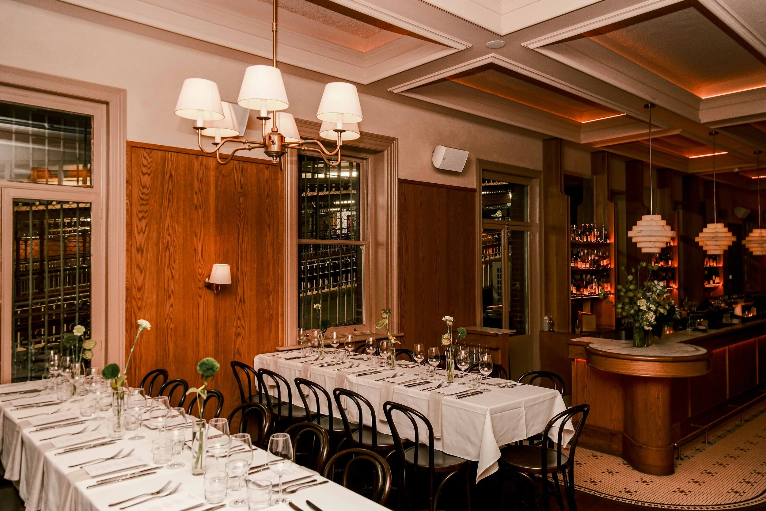 A busy bar inside a restaurant with warm lighting, pendant chandeliers, and a group of people socializing and enjoying drinks.