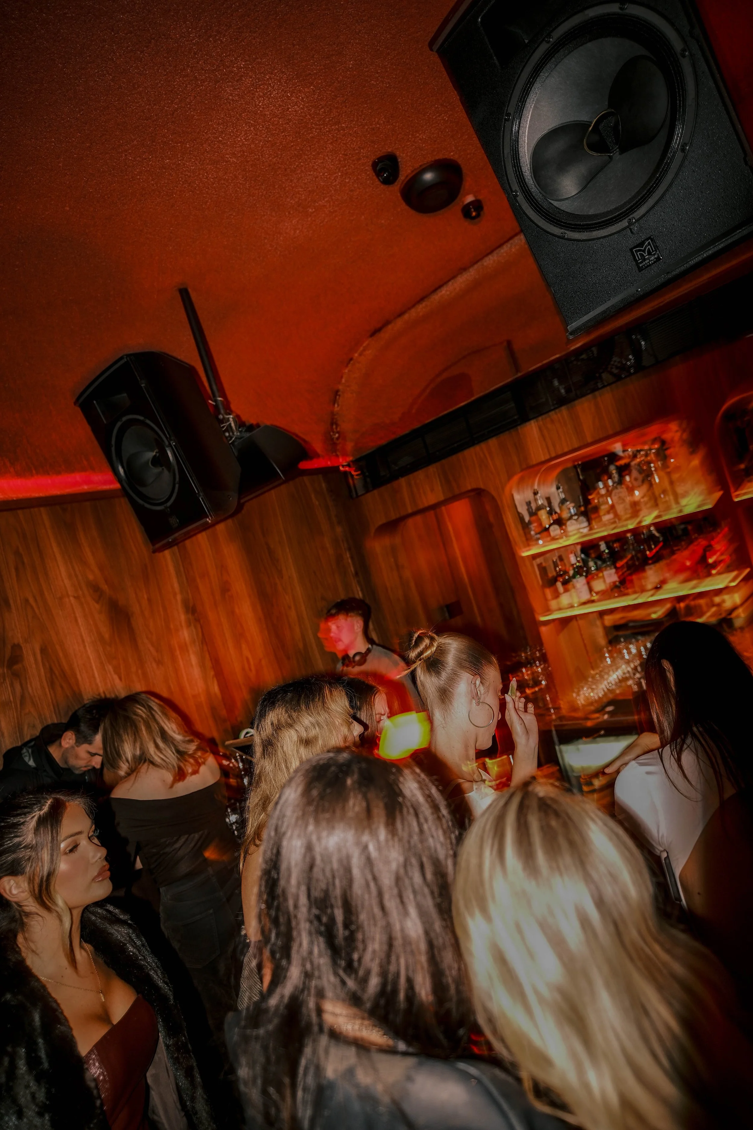 People gathered inside a bar or nightclub, watching a television screen, with a woman in a black top and denim skirt in the foreground.