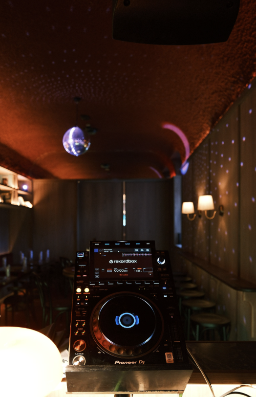 A DJ controller on a table in a dimly lit room with a red ceiling, disco balls, and wall sconces, suggesting a nightclub or party setting.