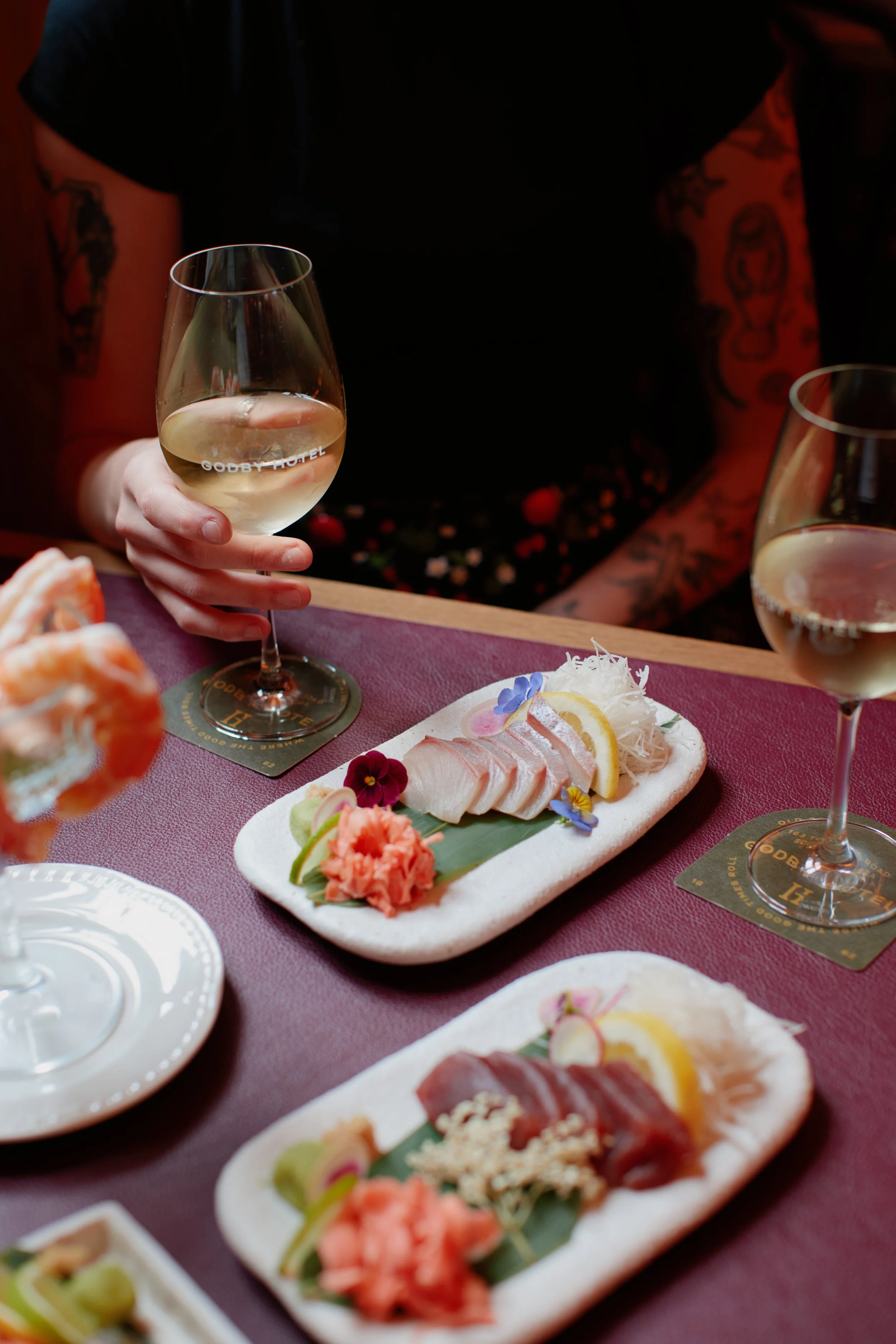 Person holding a glass of white wine at a table with plates of sashimi, garnishes, and another glass of white wine.