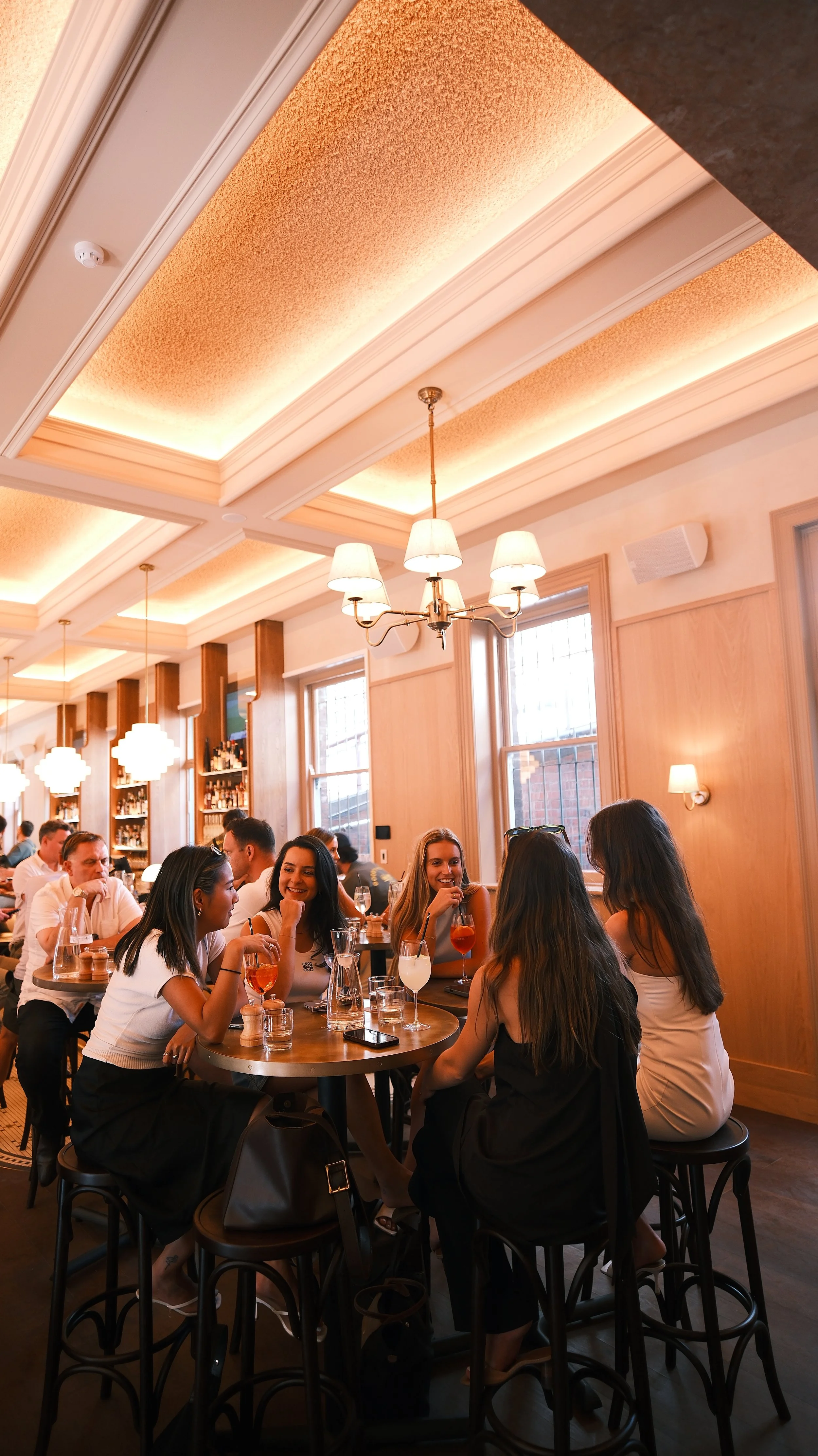 Group of women sitting around a table in a restaurant, engaging in conversation, with drinks and glasses on the table.