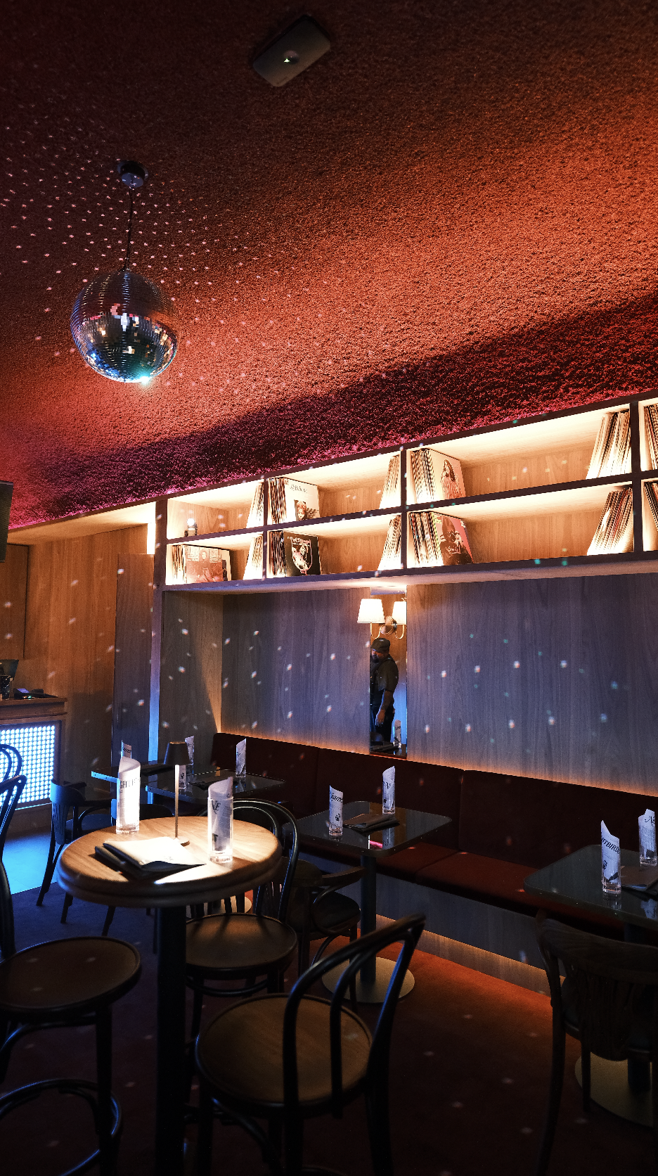 Interior of a bar or lounge with dim lighting, a disco ball hanging from the ceiling, decorated with small reflective dots, wooden shelves with books and decorative items, a mirror reflecting a staff member, and tables set with menus and water bottles.